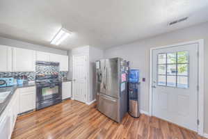 Kitchen with black range with electric cooktop, stainless steel fridge with ice dispenser, white cabinets, decorative backsplash, and light wood finished floors