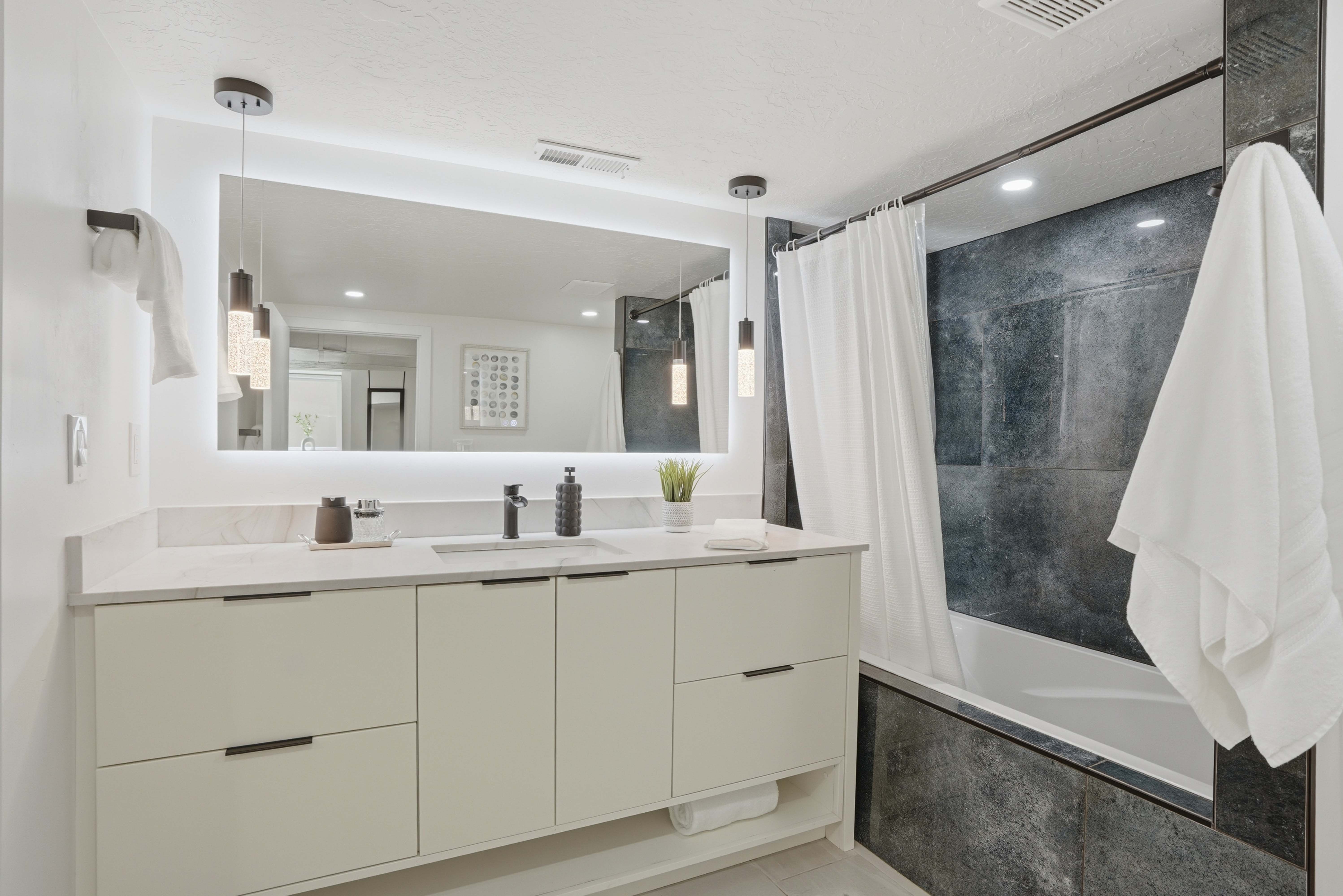Spa like bathroom featuring vanity with back light, tiled shower / bath combo, and quartzite countertops.