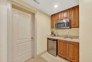 Built-in wet bar featuring wood-finish cabinetry, stainless steel microwave, stainless steel compact refrigerator, and a single basin sink set in a light-toned countertop