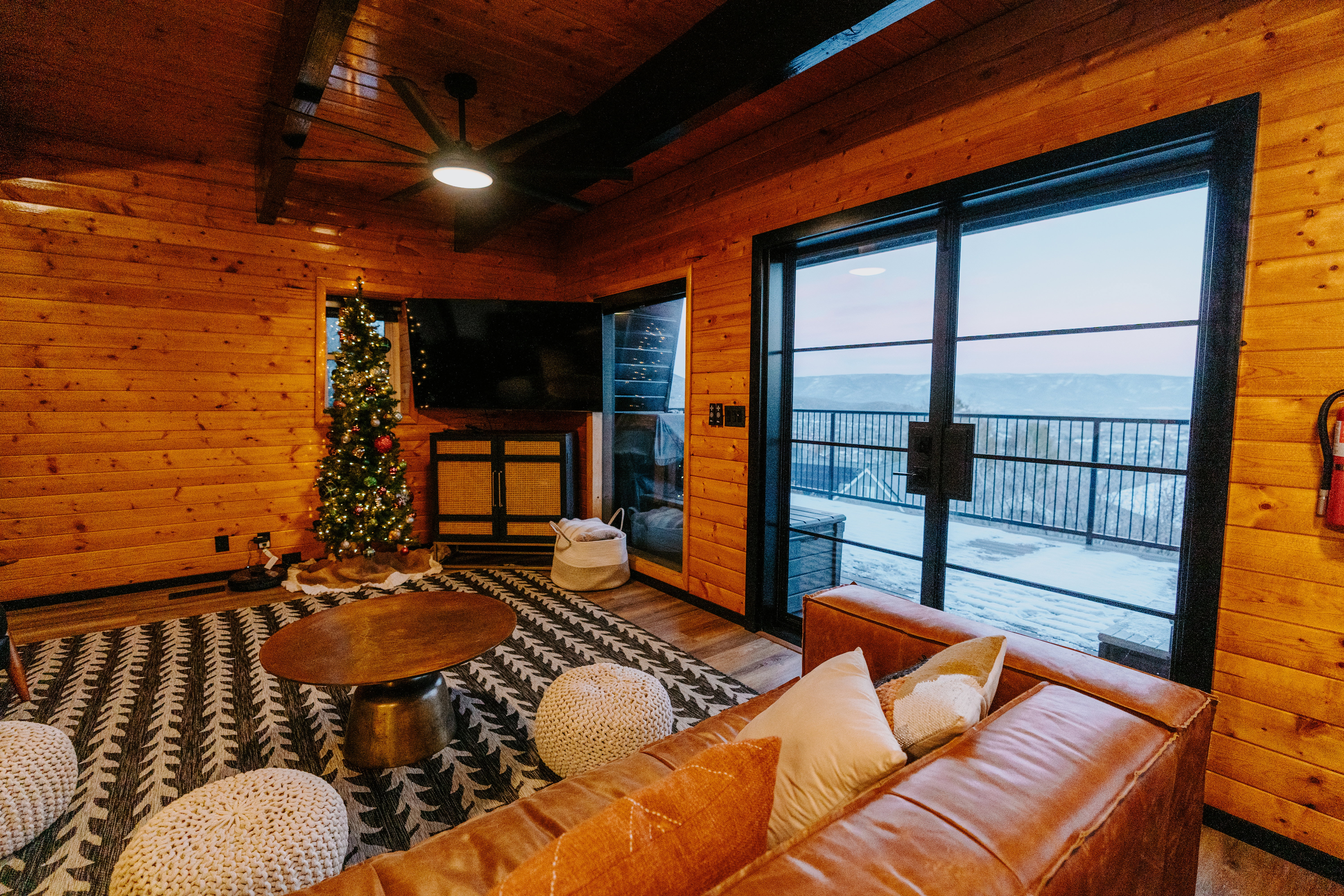 Living room with wood walls, wood finished floors, a ceiling fan, a wood ceiling with exposed beams, and a fireplace