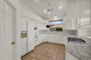 Kitchen featuring tile countertops, white appliances, vaulted ceiling, white cabinetry, and tasteful backsplash