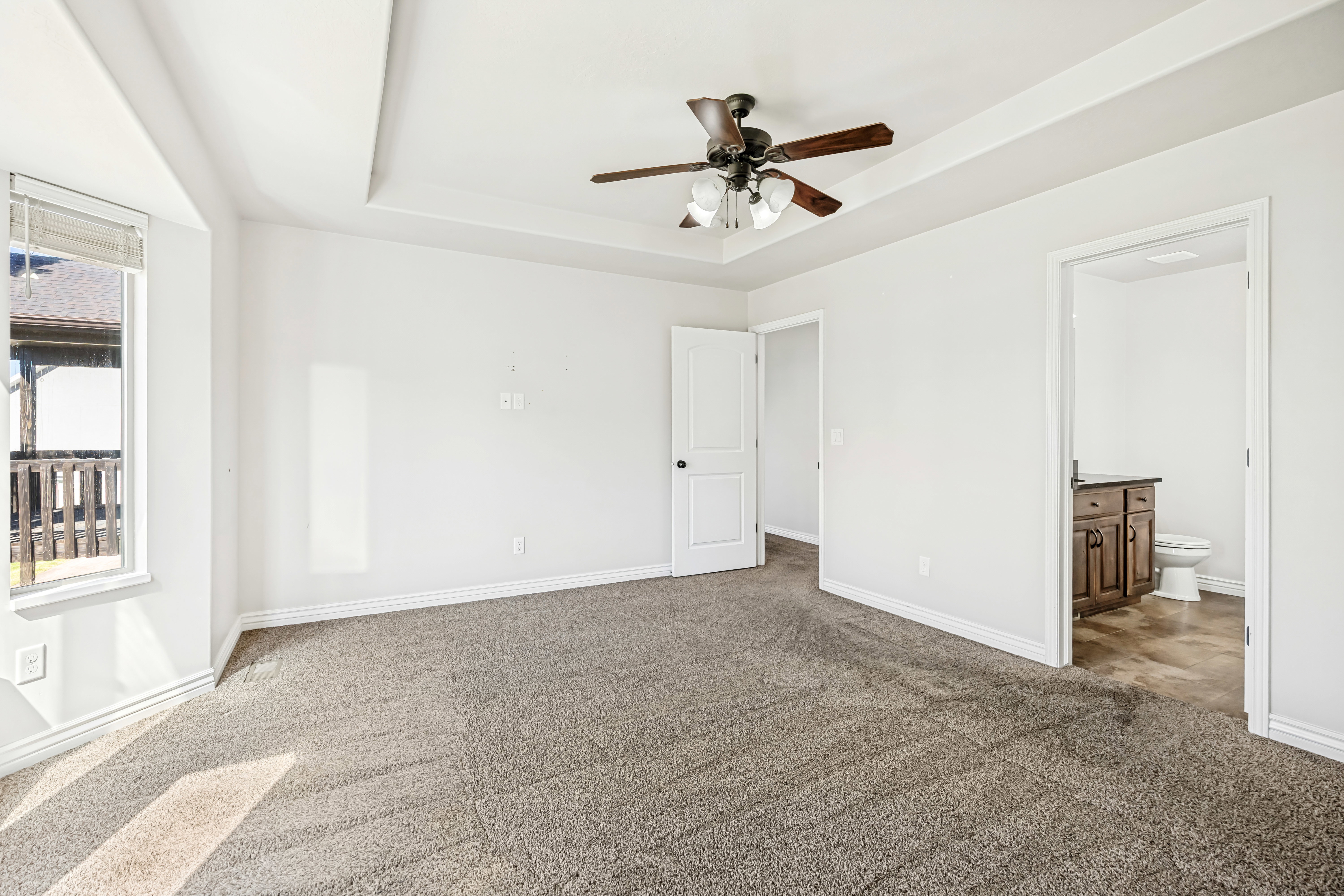 Unfurnished bedroom with dark colored carpet, a raised ceiling, connected bathroom, and ceiling fan