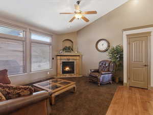 Living area featuring a corner fireplace with stone surround and mantle, vaulted ceilings, a ceiling fan, and multiple windows with horizontal blinds