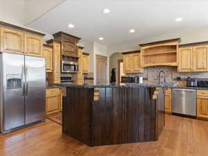 Kitchen featuring an expansive dark wood-paneled island with an integrated sink, complementing light wood-finish cabinetry, stainless steel appliances, a tiled backsplash, and wood-finish flooring