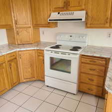 Kitchen with white electric range oven, range hood, wood finish cabinetry, and light tile patterned floors
