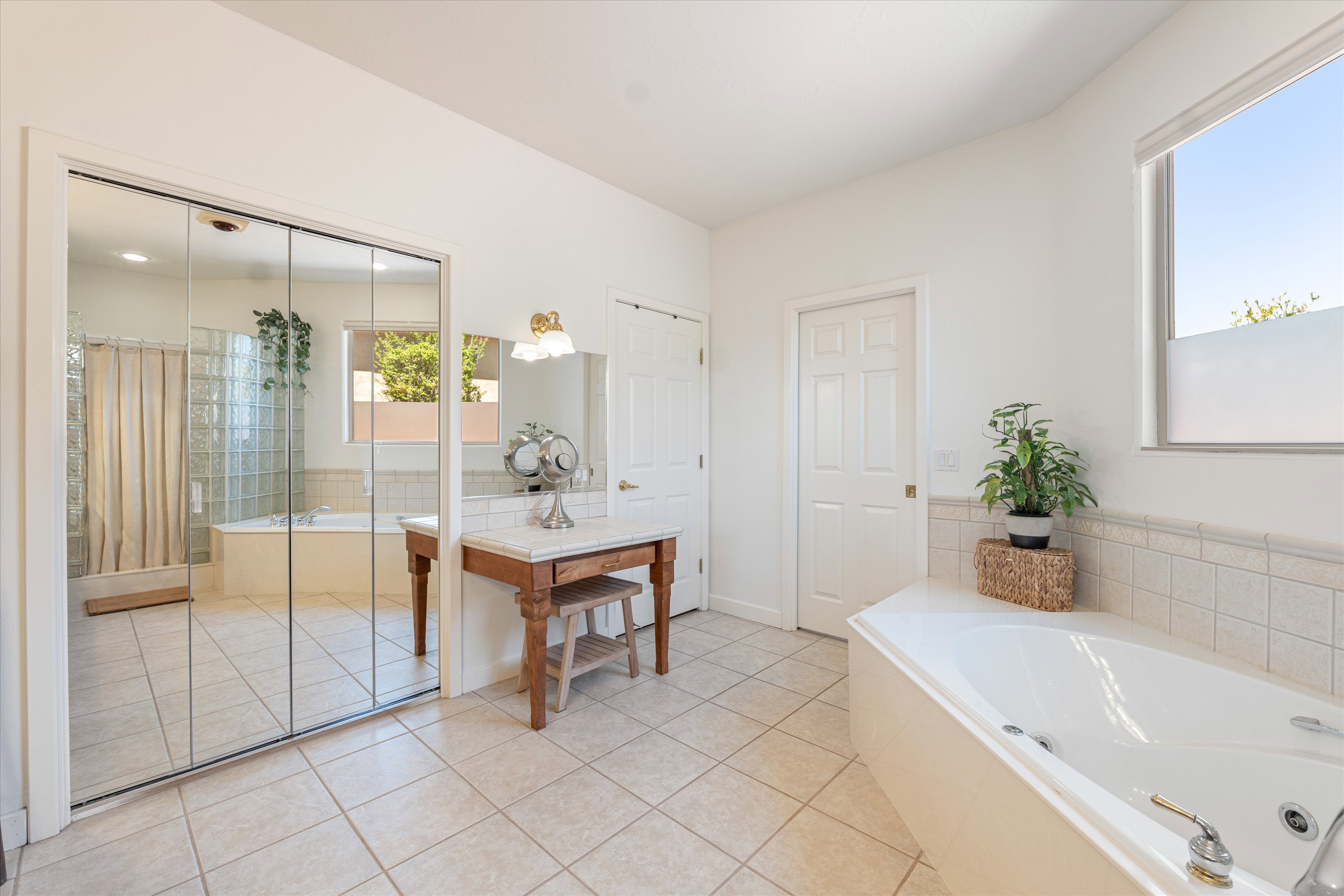 Spacious bathroom featuring a white soaking tub, tiled flooring, mirrored closet doors, a vanity with a light-colored countertop, and a large window providing natural illumination