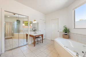Spacious bathroom featuring a white soaking tub, tiled flooring, mirrored closet doors, a vanity with a light-colored countertop, and a large window providing natural illumination