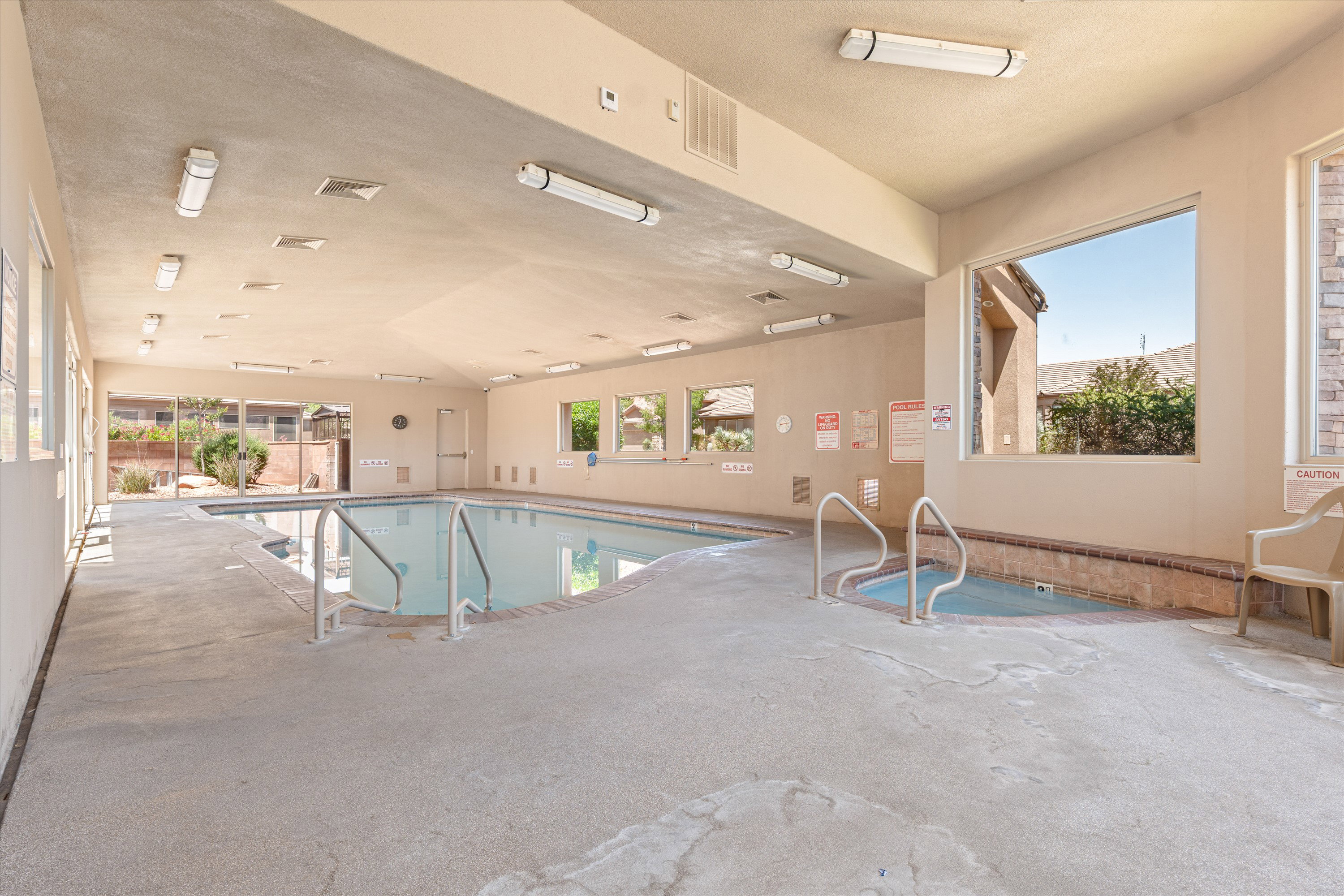 Indoor swimming pool with an integrated spa, featuring concrete decking, multiple windows, and fluorescent overhead lighting