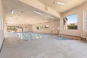 Indoor swimming pool with an integrated spa, featuring concrete decking, multiple windows, and fluorescent overhead lighting