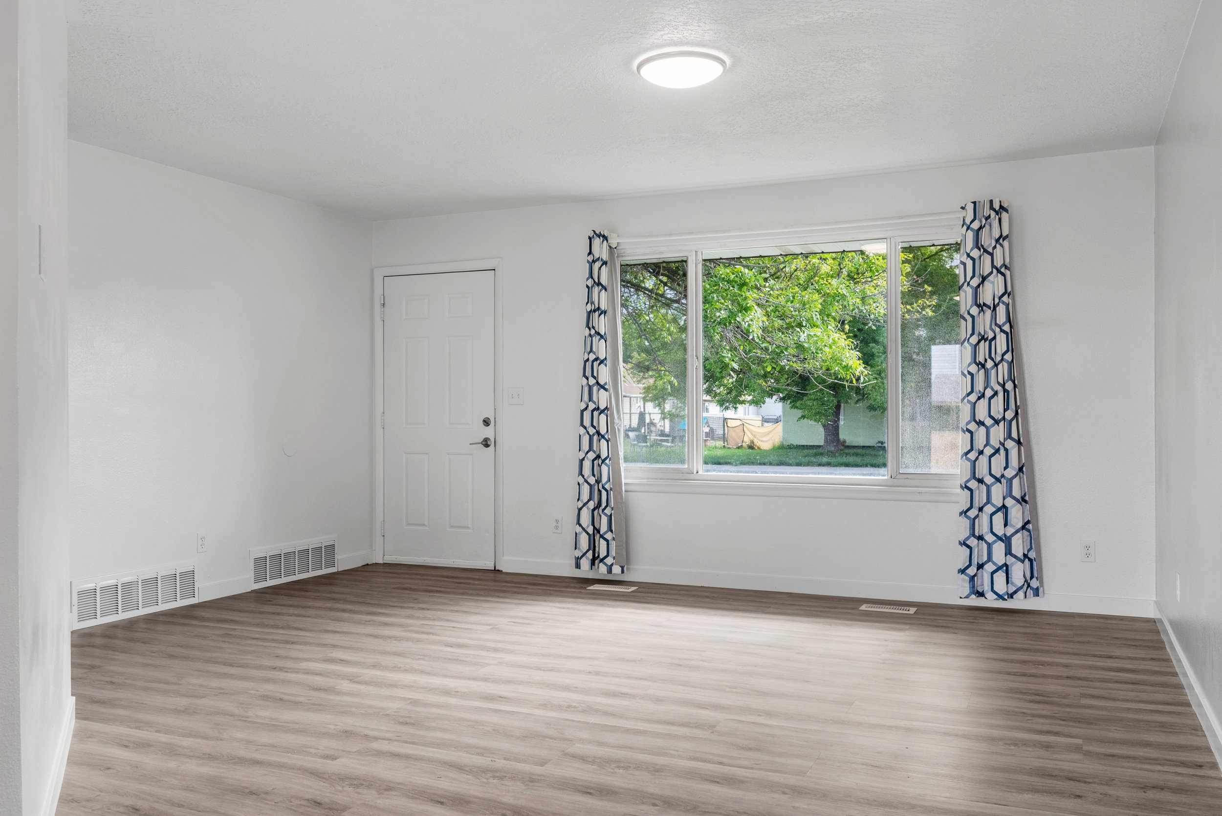 Living space featuring wood-finish flooring, white walls, and a large window with patterned drapes