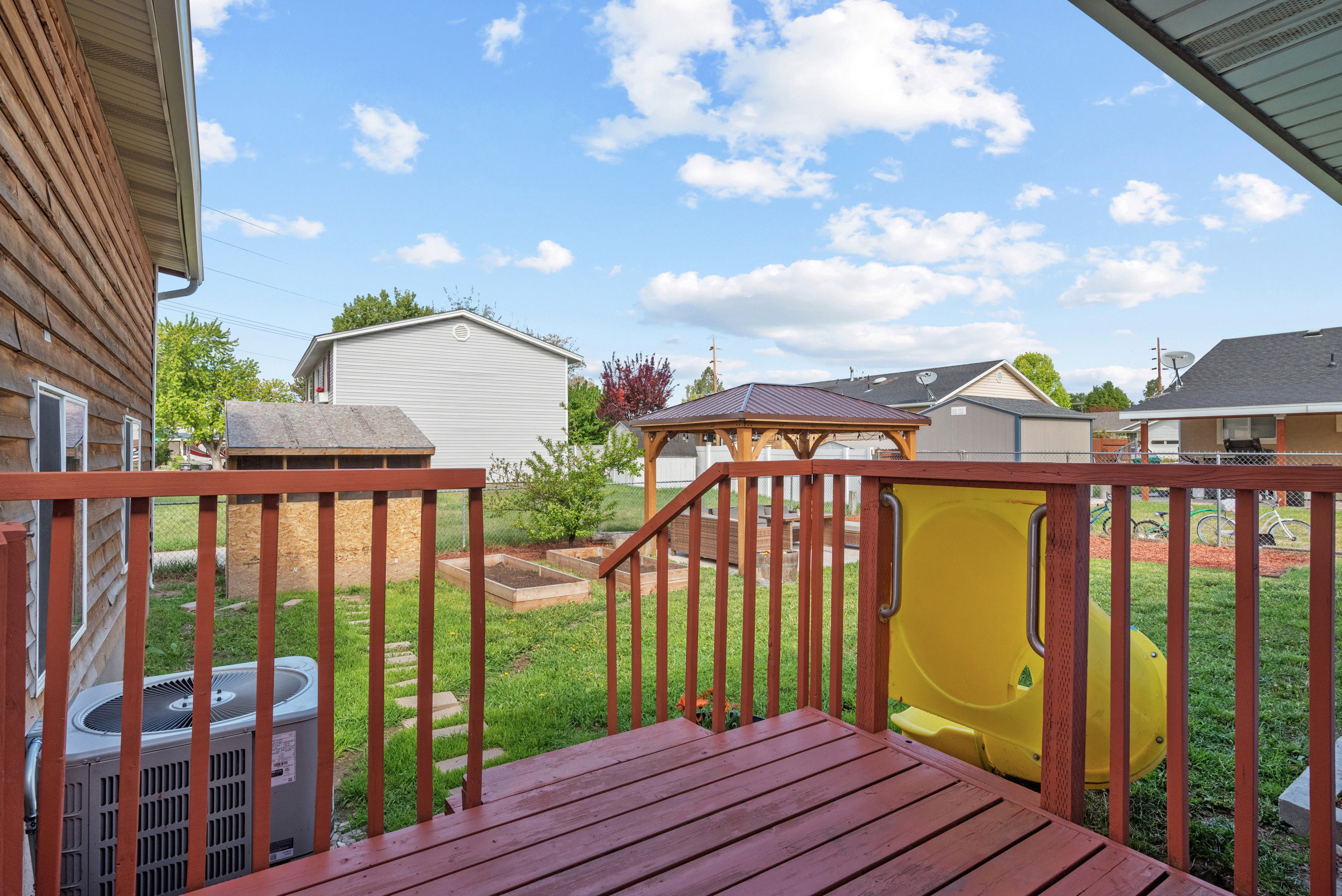 Expansive backyard featuring a wood-stained deck, a detached shed, raised garden beds, a gazebo, and a chain-link fence