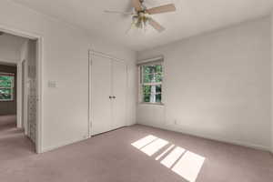Neutral tone wall paint, light-colored carpeting, a window with exterior tree foliage visible, a ceiling fan, and white bi-fold closet doors