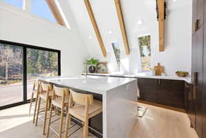 Modern kitchen featuring a waterfall island, light wood-finish flooring, dark wood cabinetry, white countertops, and exposed wood beams