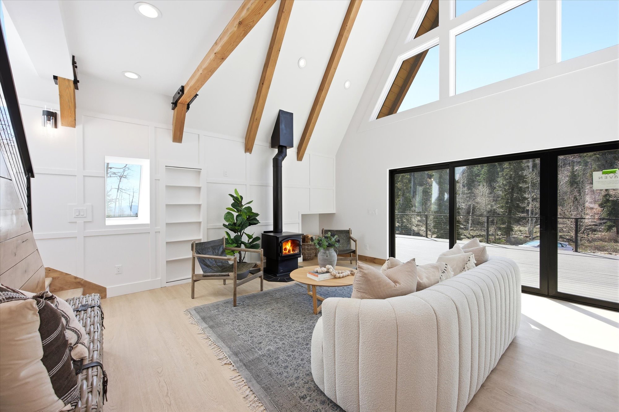Living area featuring an exposed beam vaulted ceiling, a freestanding wood-burning stove, large windows, sliding glass doors, and light wood-finish flooring