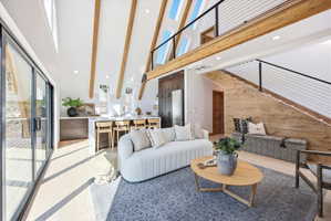 Vaulted ceilings with exposed wood beams, a modern kitchen island with seating, wide plank wood-finish flooring, a wall of glass sliding doors, and a staircase featuring a wood plank wall and cable railing