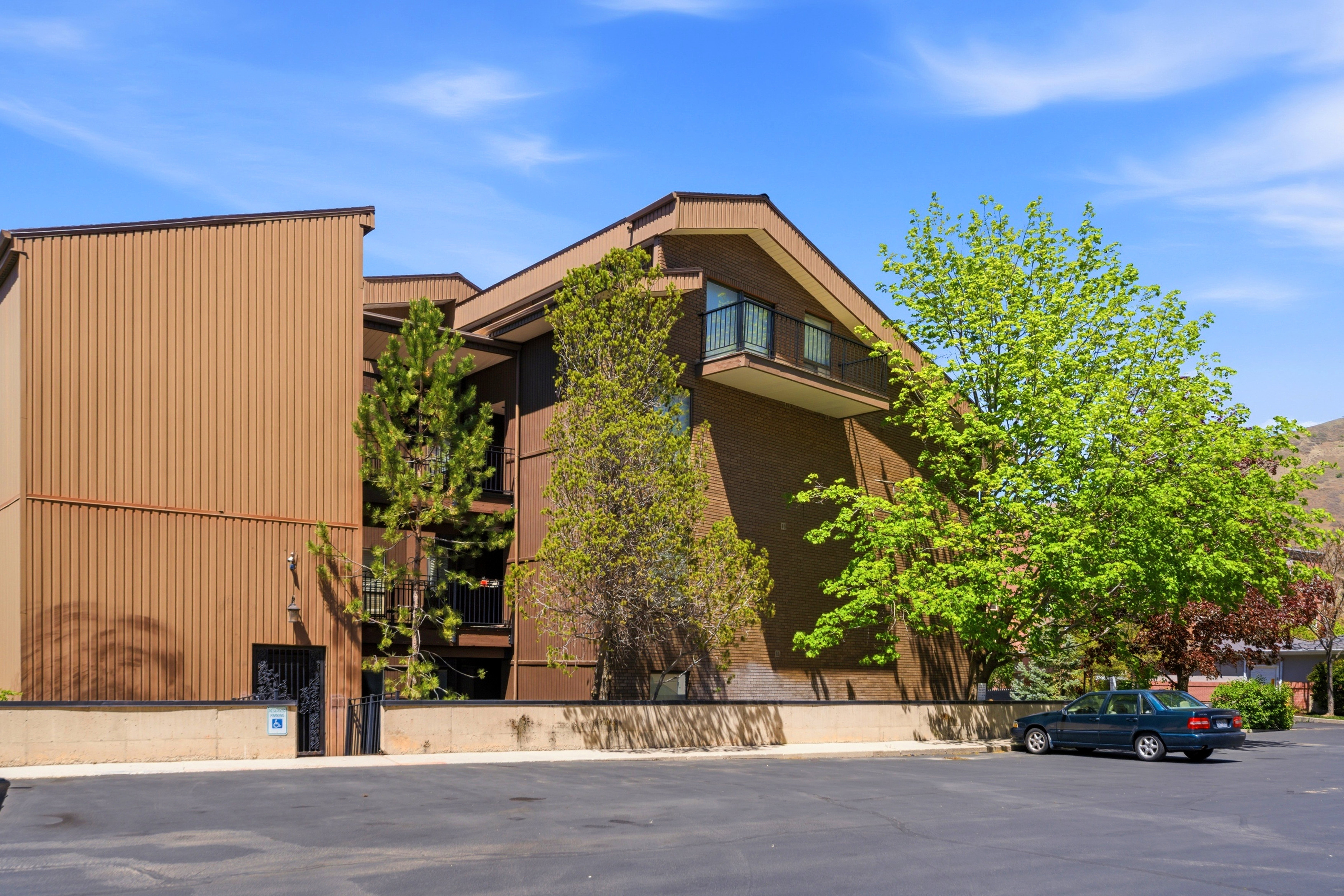 Multi-story building featuring a brown brick and corrugated metal exterior, an upper-level balcony with a black railing, and mature trees providing natural screening