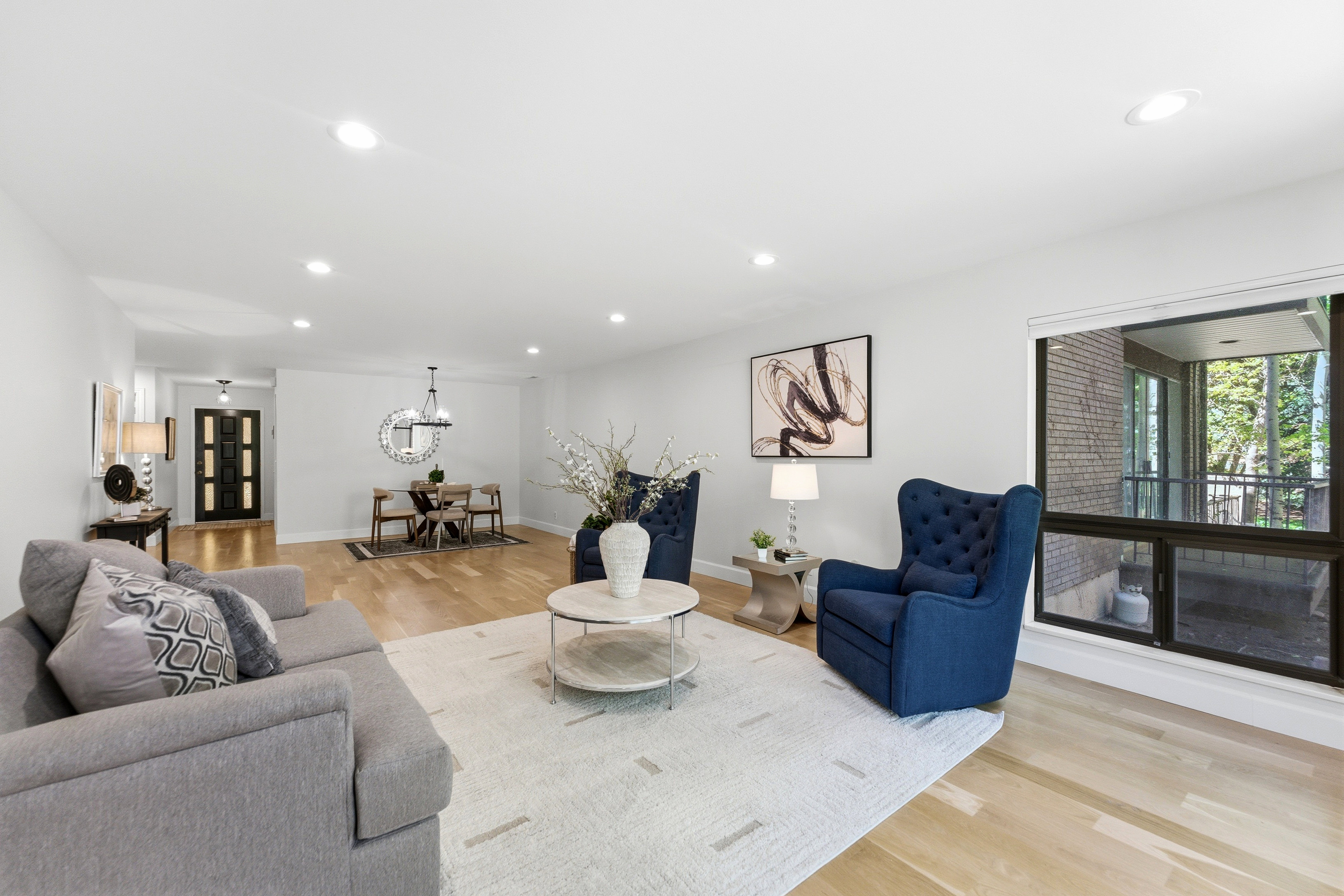 Open-concept living area featuring light wood-finish flooring, recessed lighting, a wide window, and a formal dining space with a contemporary chandelier
