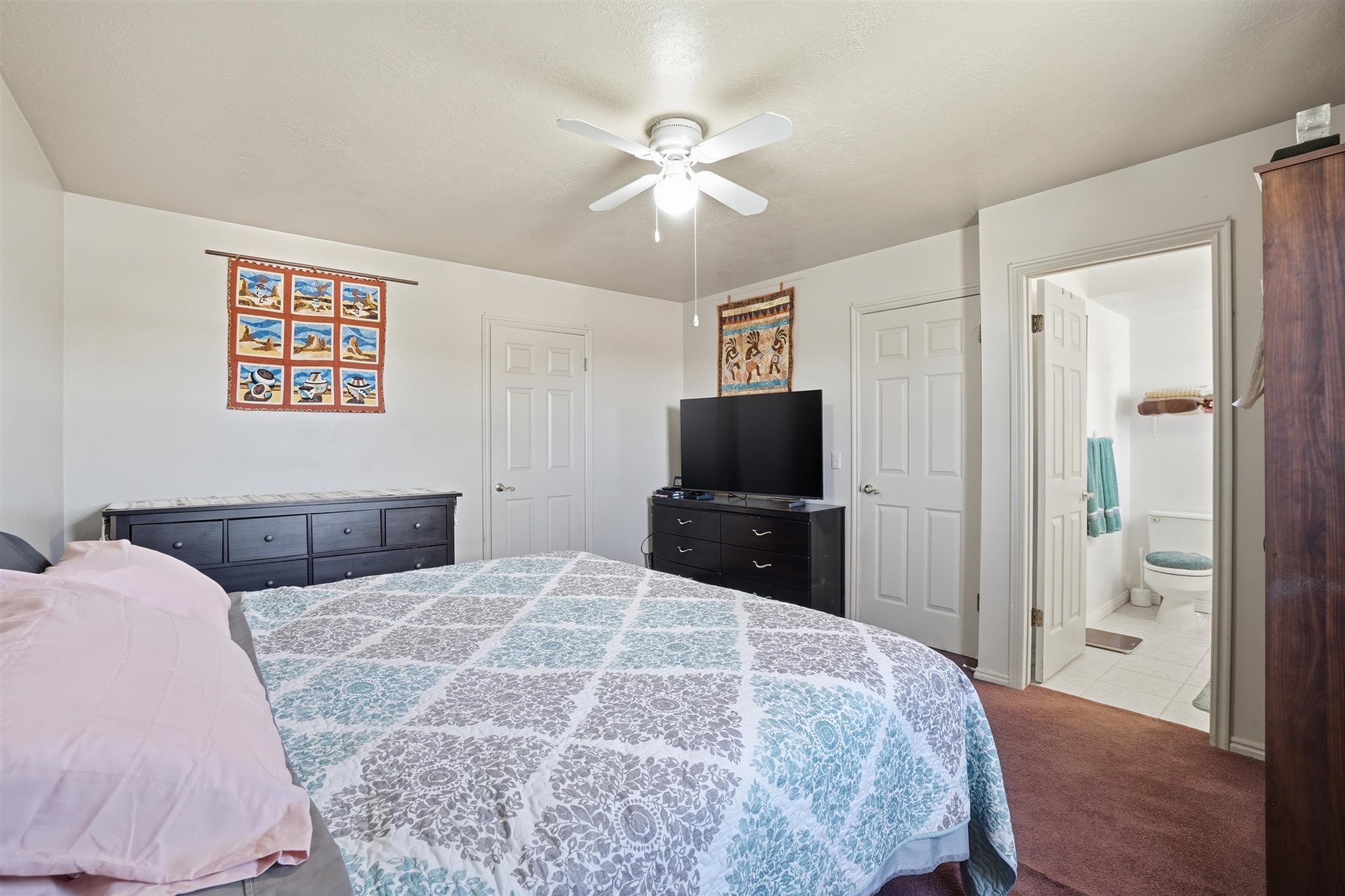 Carpeted bedroom featuring a ceiling fan with light fixture, white interior doors, and an open doorway to a bathroom with tile flooring and a toilet