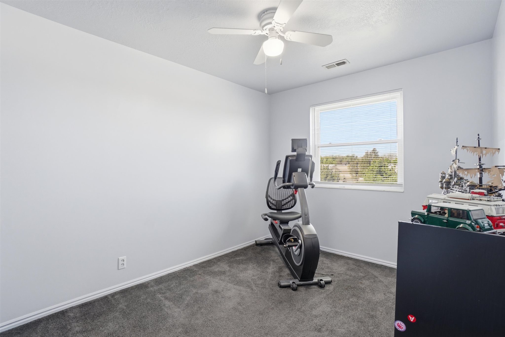 Bright room featuring a window with blinds, ceiling fan with light, gray wall-to-wall carpeting, and white baseboards