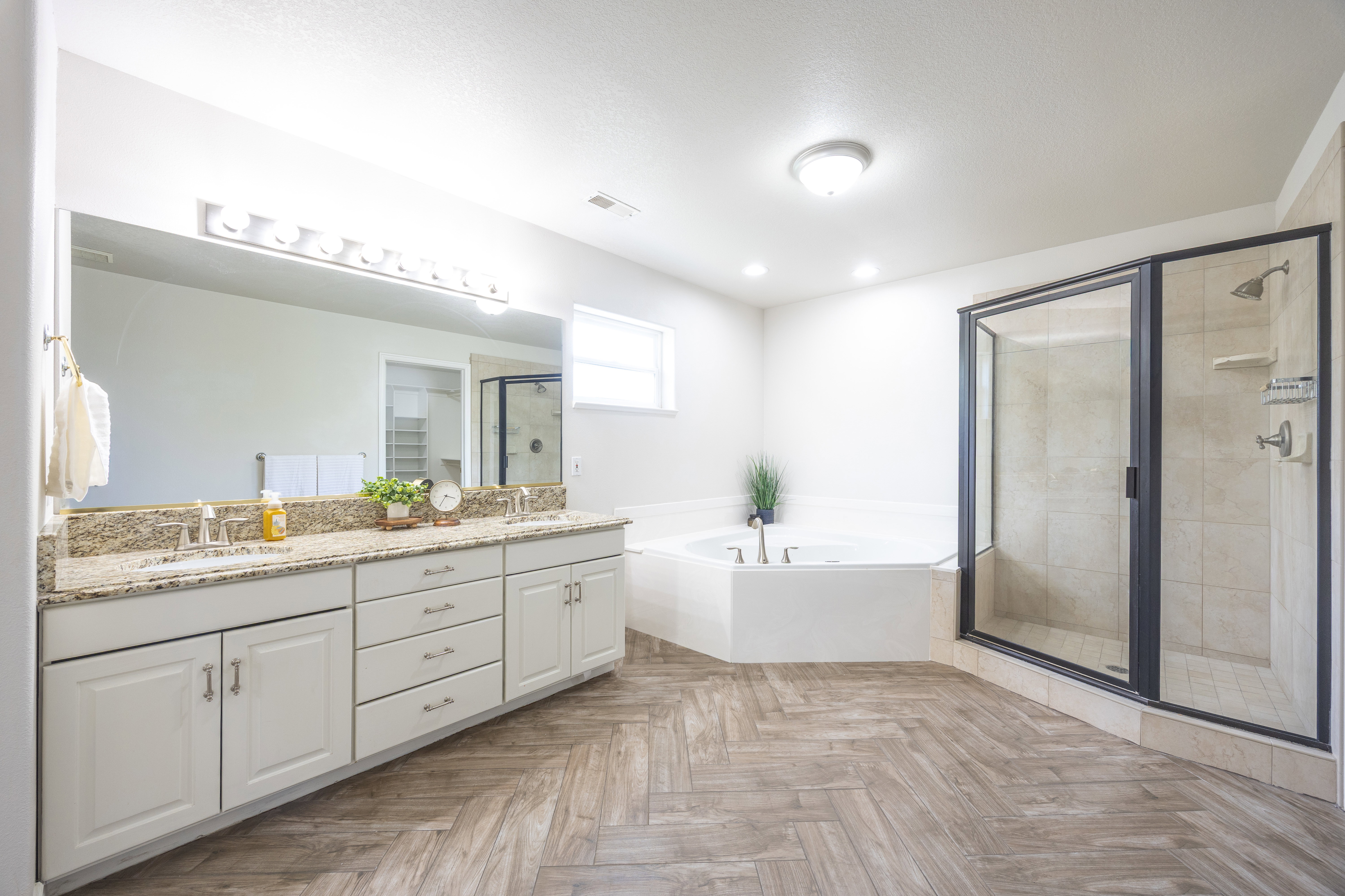 Spacious bathroom featuring a dual vanity with a granite countertop, a large walk-in shower with a glass enclosure, a corner soaking tub, and wood-finish flooring