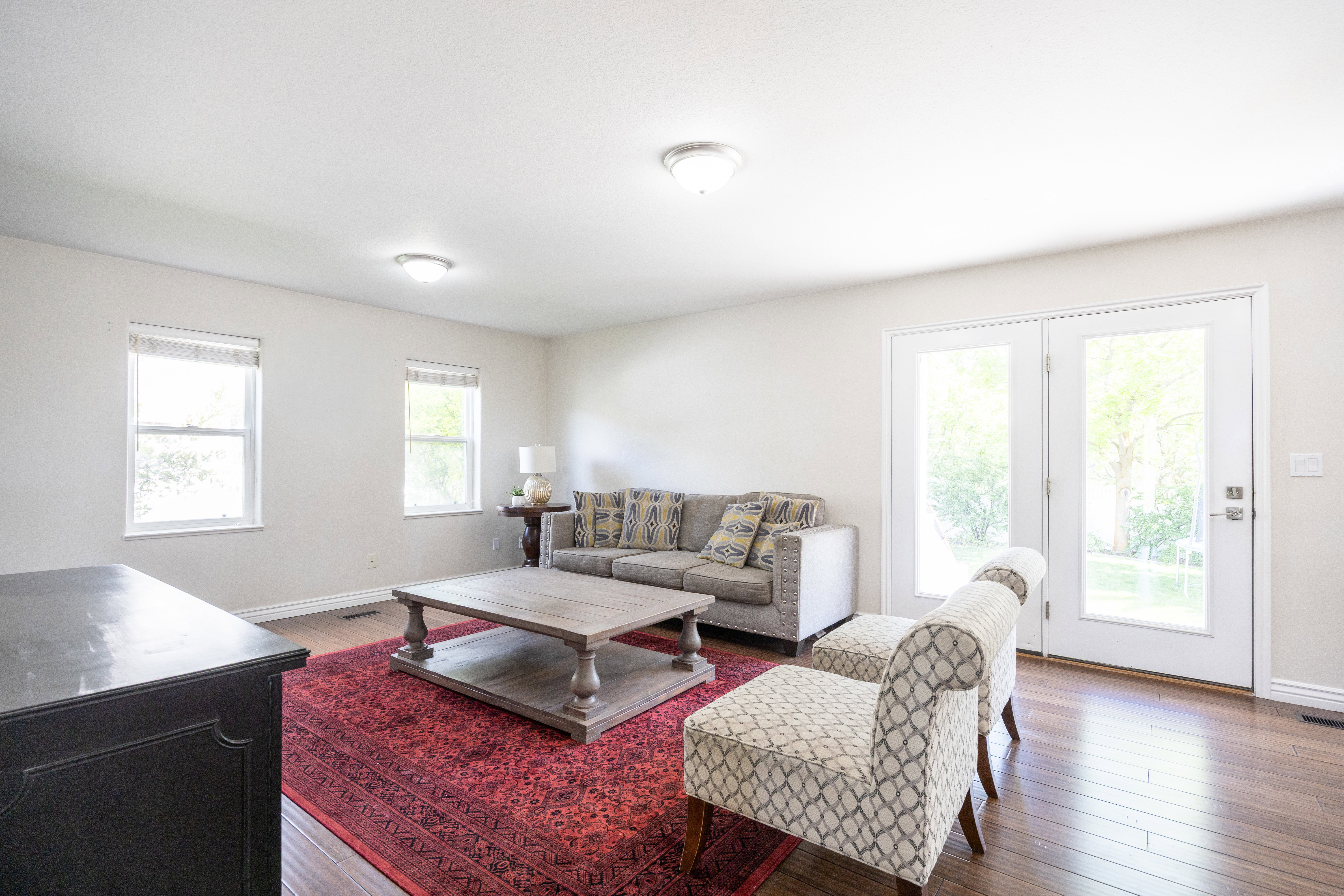 Living area featuring wood-finish flooring, light neutral wall tones, two windows with blinds, two ceiling-mounted light fixtures, and white French doors
