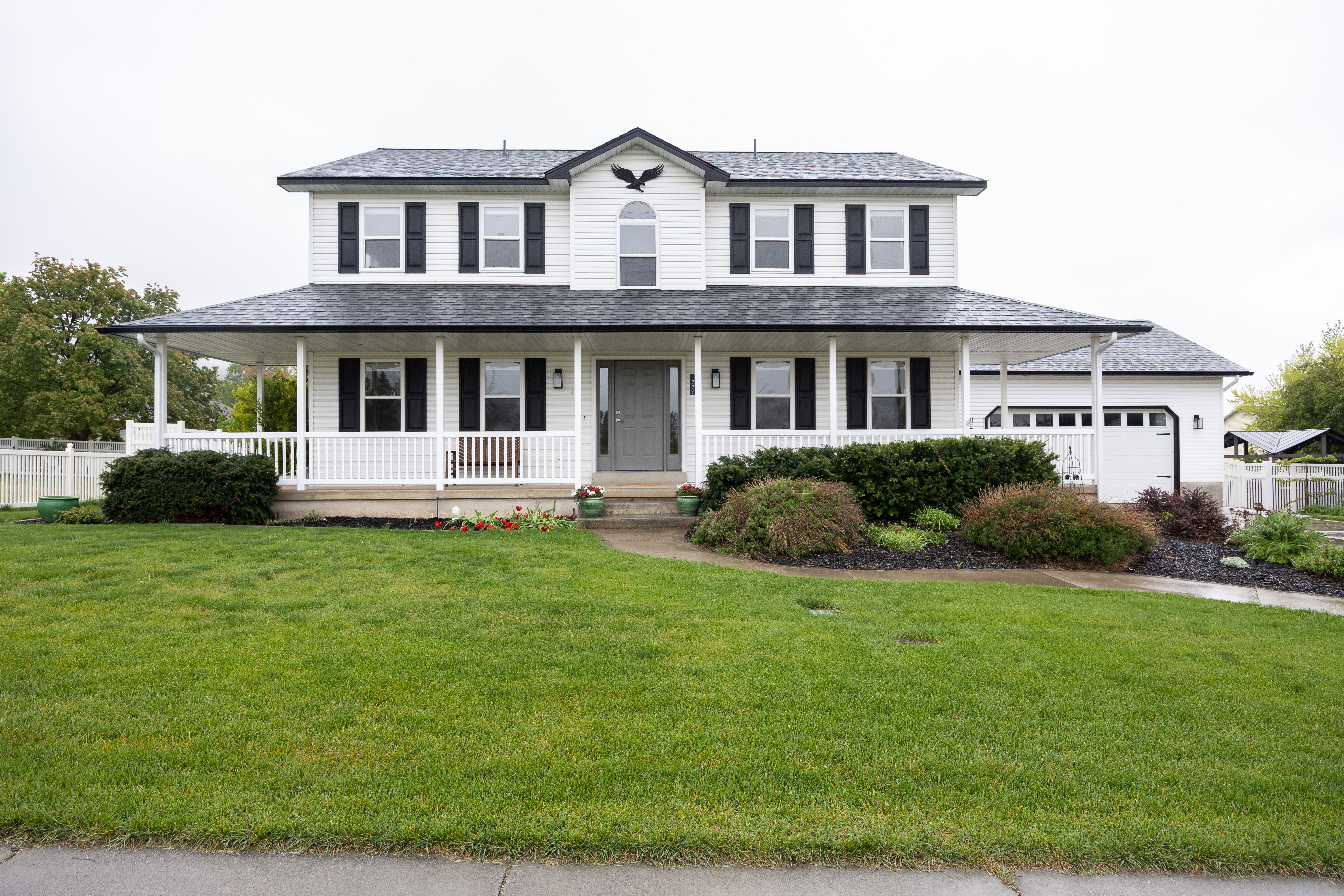 Colonial-style residence featuring a prominent covered front porch, white siding, black window shutters, a two-car garage, and established landscaping