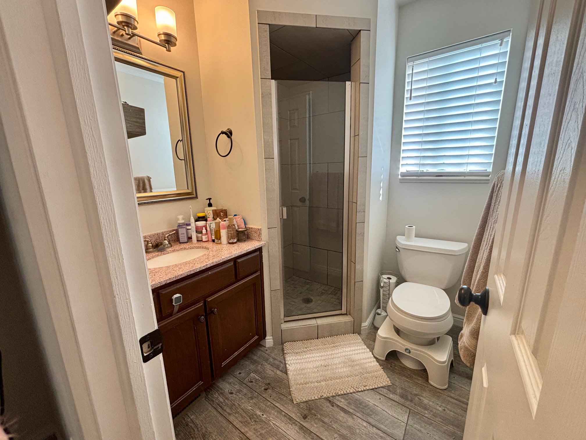 Bathroom featuring a wood-finish vanity with a built-in sink and stone countertop, a glass-enclosed shower with tile surround, a toilet, a window with horizontal blinds, and wood-finish flooring