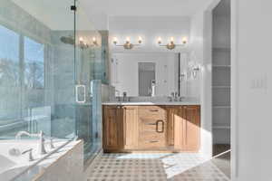 Bathroom featuring a glass-enclosed shower with a rainfall showerhead, a built-in soaking tub, a dual vanity with a light wood-finish cabinet and white countertop, and patterned floor tiling