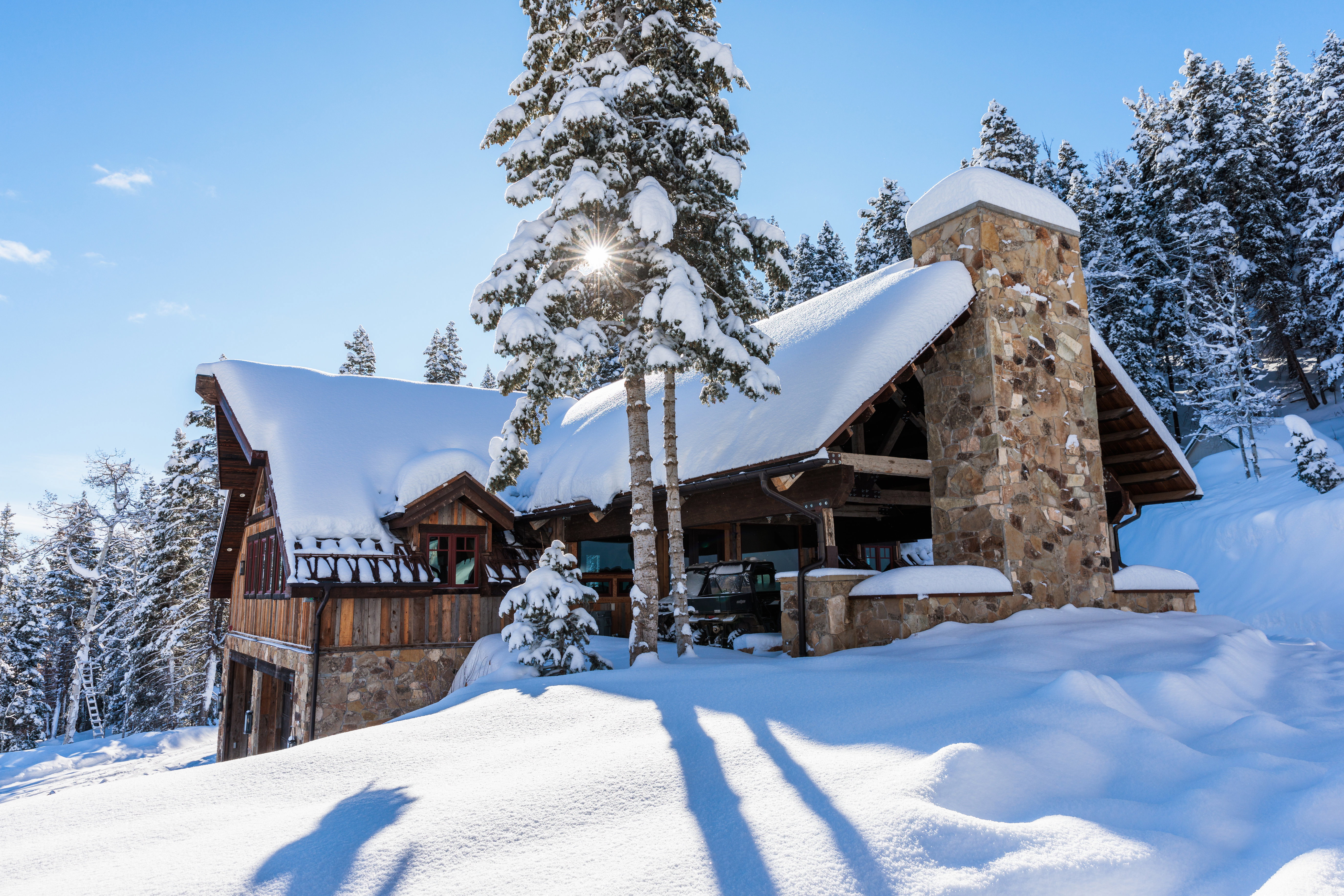 Stone and wood-sided exterior featuring a prominent stone chimney, a covered outdoor area with exposed beams, and a snow-covered roofline