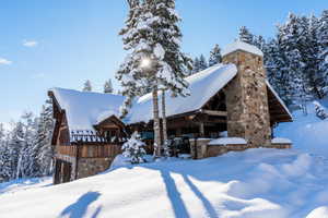 Stone and wood-sided exterior featuring a prominent stone chimney, a covered outdoor area with exposed beams, and a snow-covered roofline