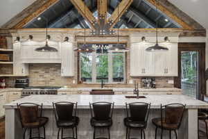 Spacious kitchen featuring a large island with seating, a professional-grade range, light-toned cabinetry, exposed brick backsplash, and architectural ceiling beams