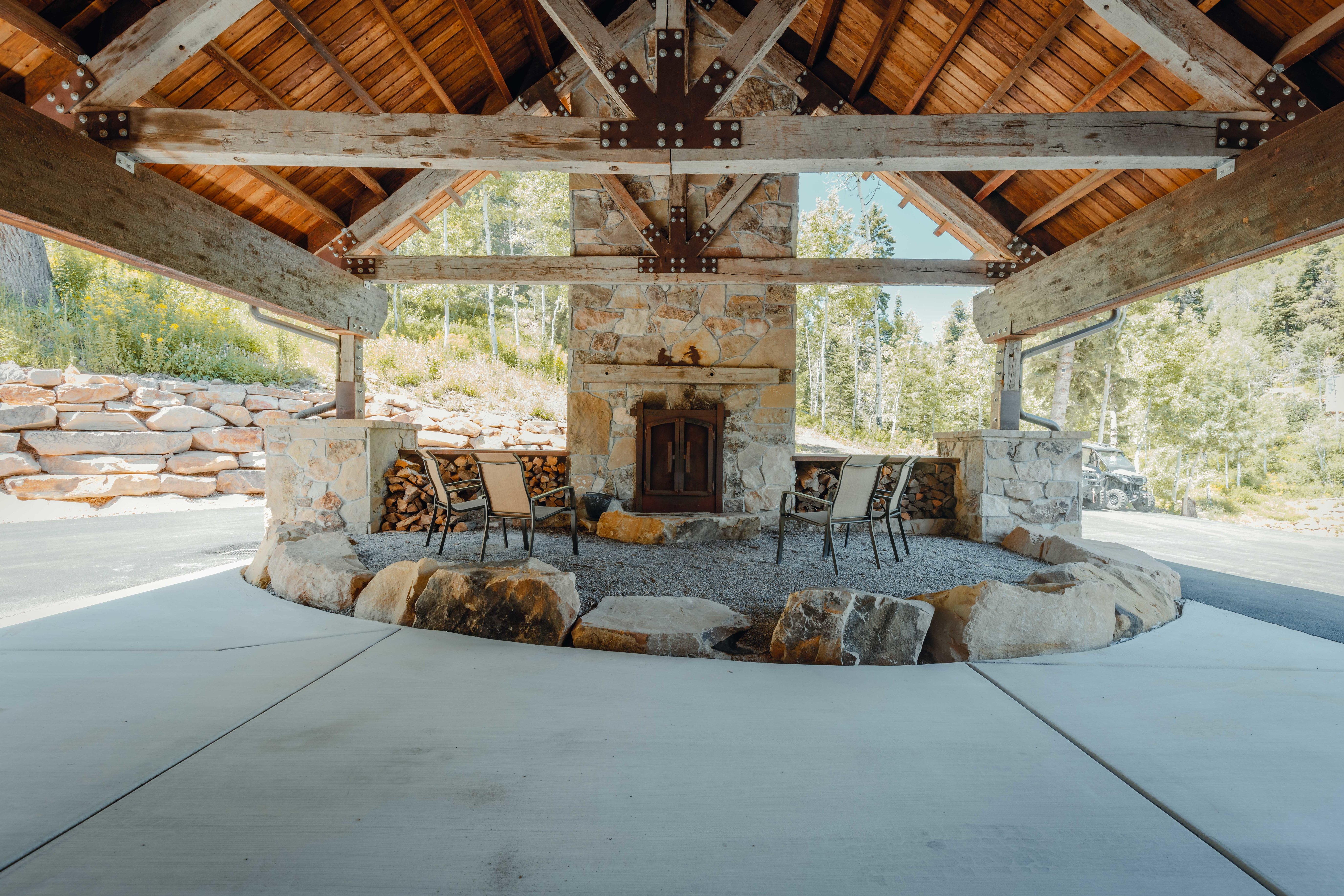 Expansive outdoor covered patio featuring a stone fireplace, exposed timber beams, Tongue and Groove ceiling, concrete flooring, and natural boulder accents