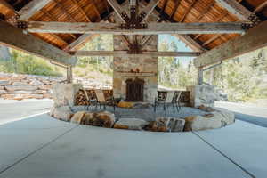Expansive outdoor covered patio featuring a stone fireplace, exposed timber beams, Tongue and Groove ceiling, concrete flooring, and natural boulder accents