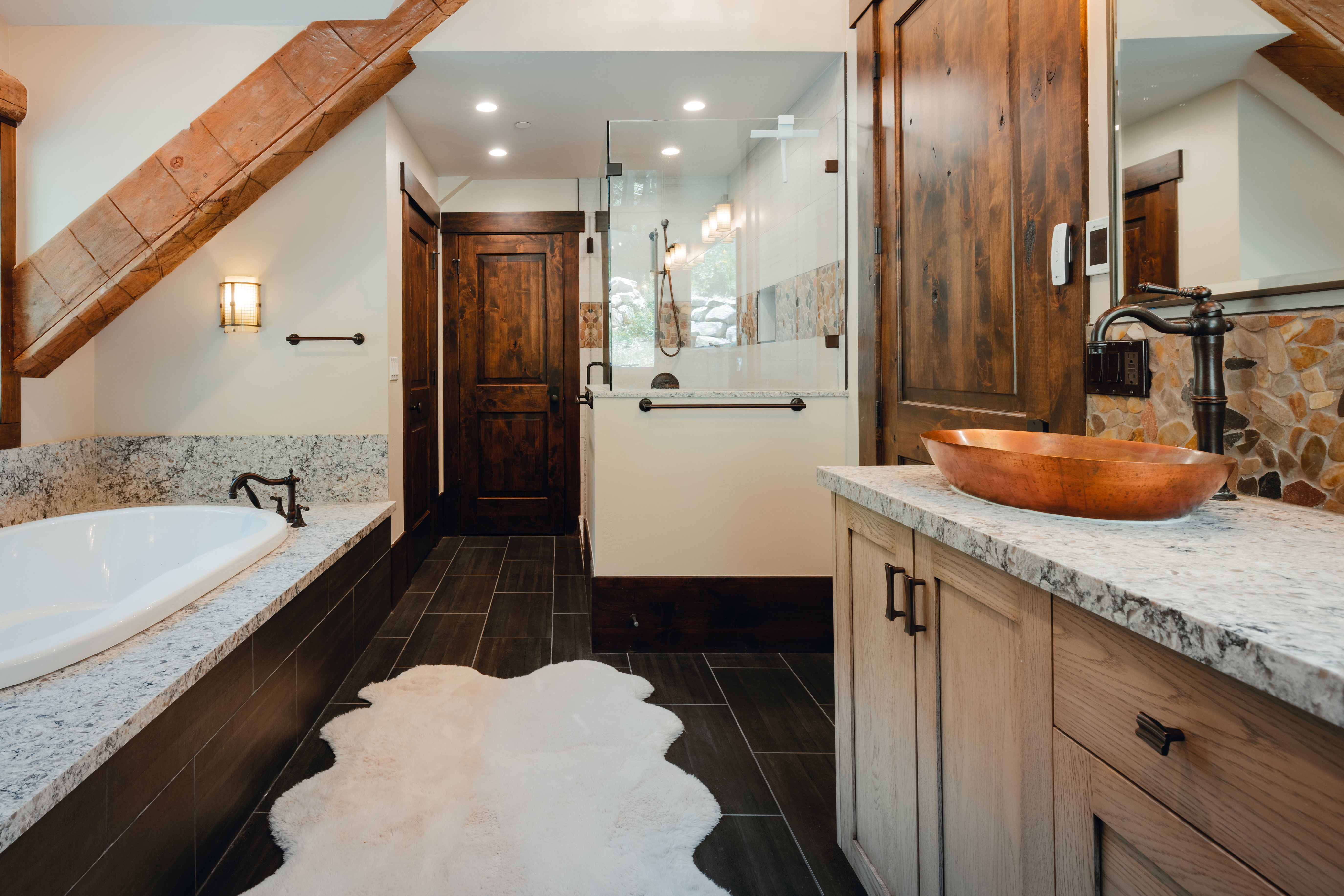 Bathroom featuring a full-size soaking tub with a stone-finish surround, a frameless glass shower enclosure, a vanity with a copper vessel sink and stone countertop, wood-finish flooring, and exposed ceiling beams