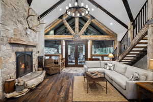 Vaulted ceiling great room featuring exposed timber beams, a stone fireplace with a substantial hearth, wood-finish flooring, and a large window wall with exterior doors