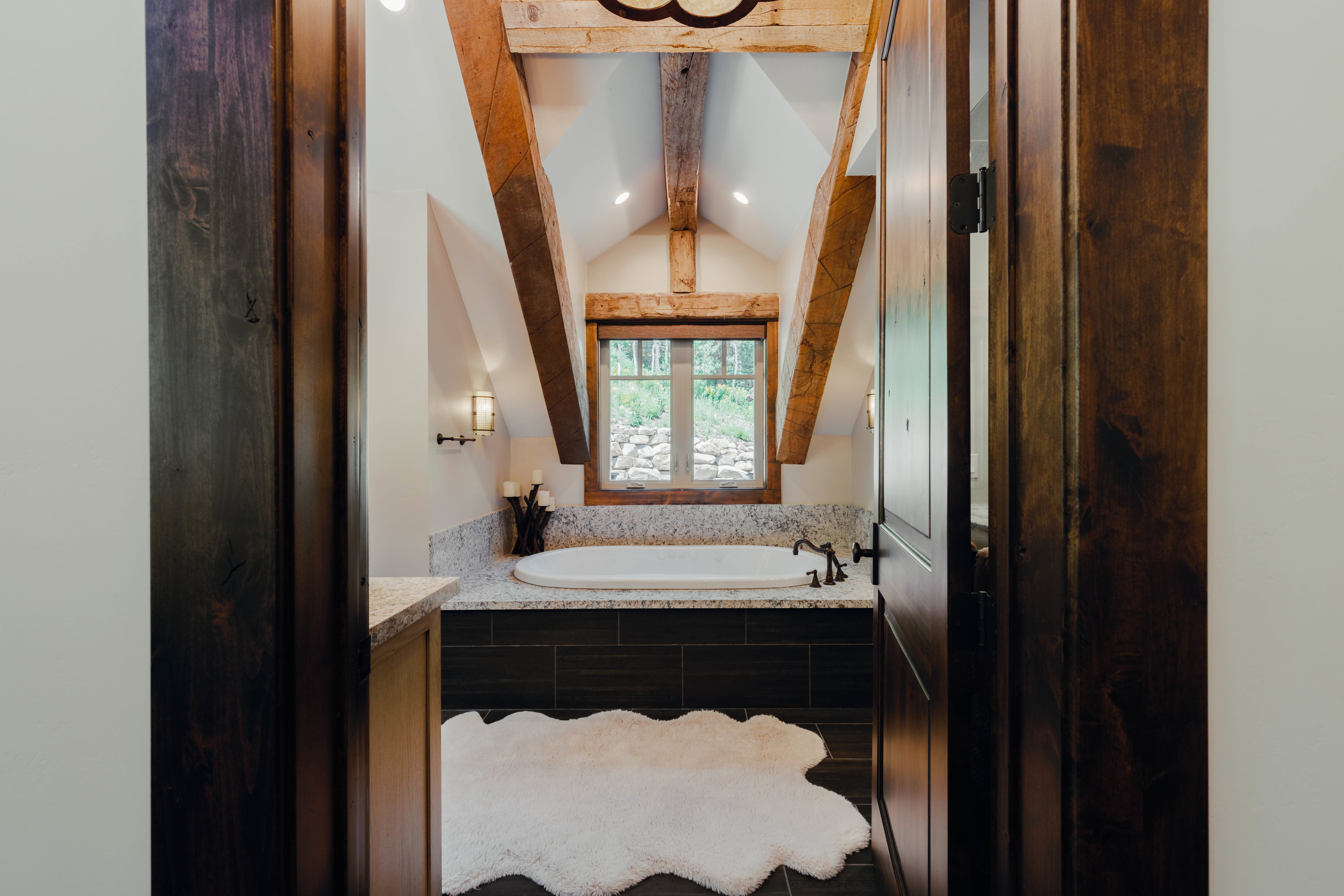 Attic bathroom featuring exposed wood beams, a built-in soaking tub with a dark tile surround, a wood-framed window, recessed lighting, and sconce lighting