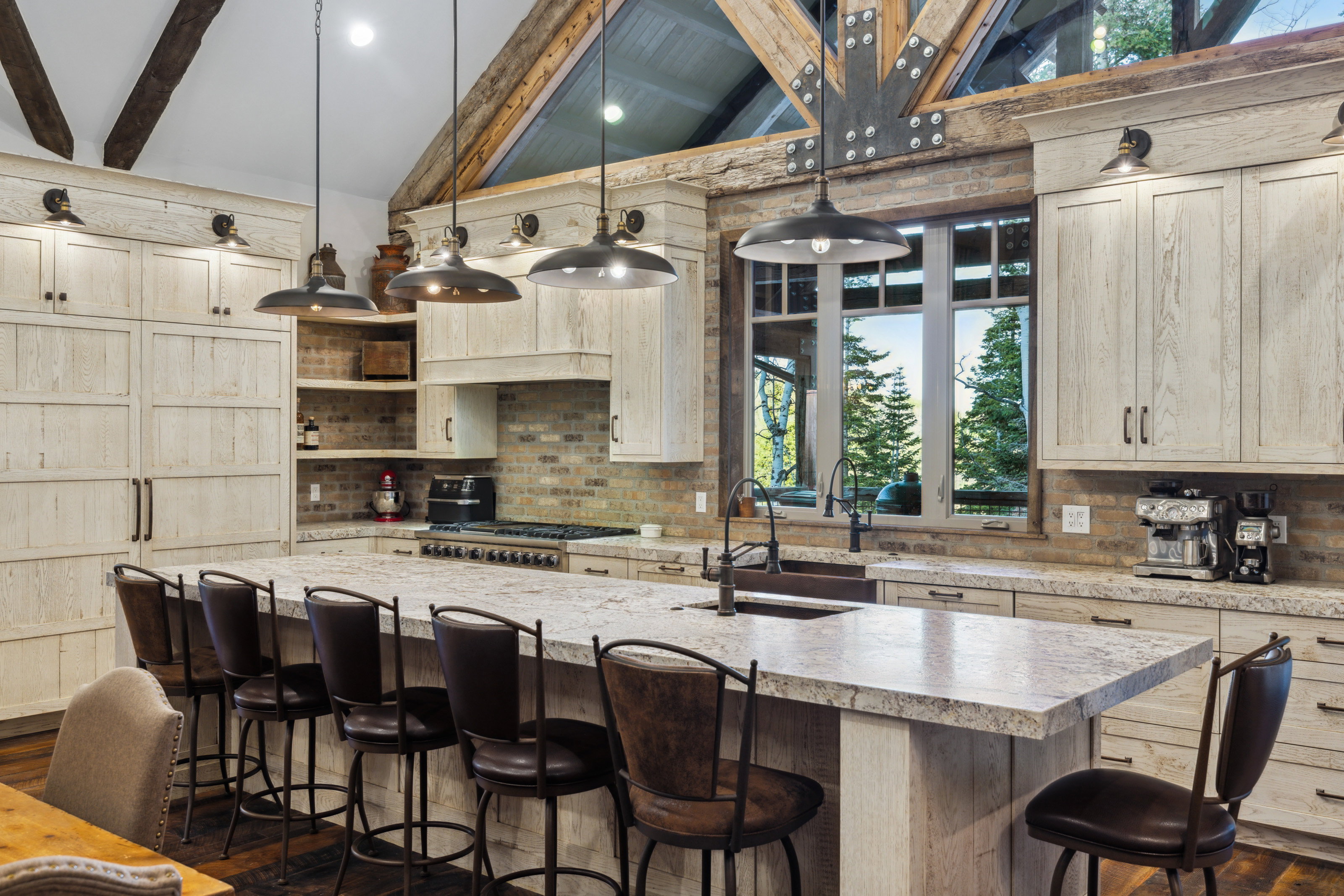 Expansive kitchen featuring a large island with countertop seating, stone countertops, a full-height brick backsplash, exposed wood beam ceiling, and multiple pendant light fixtures