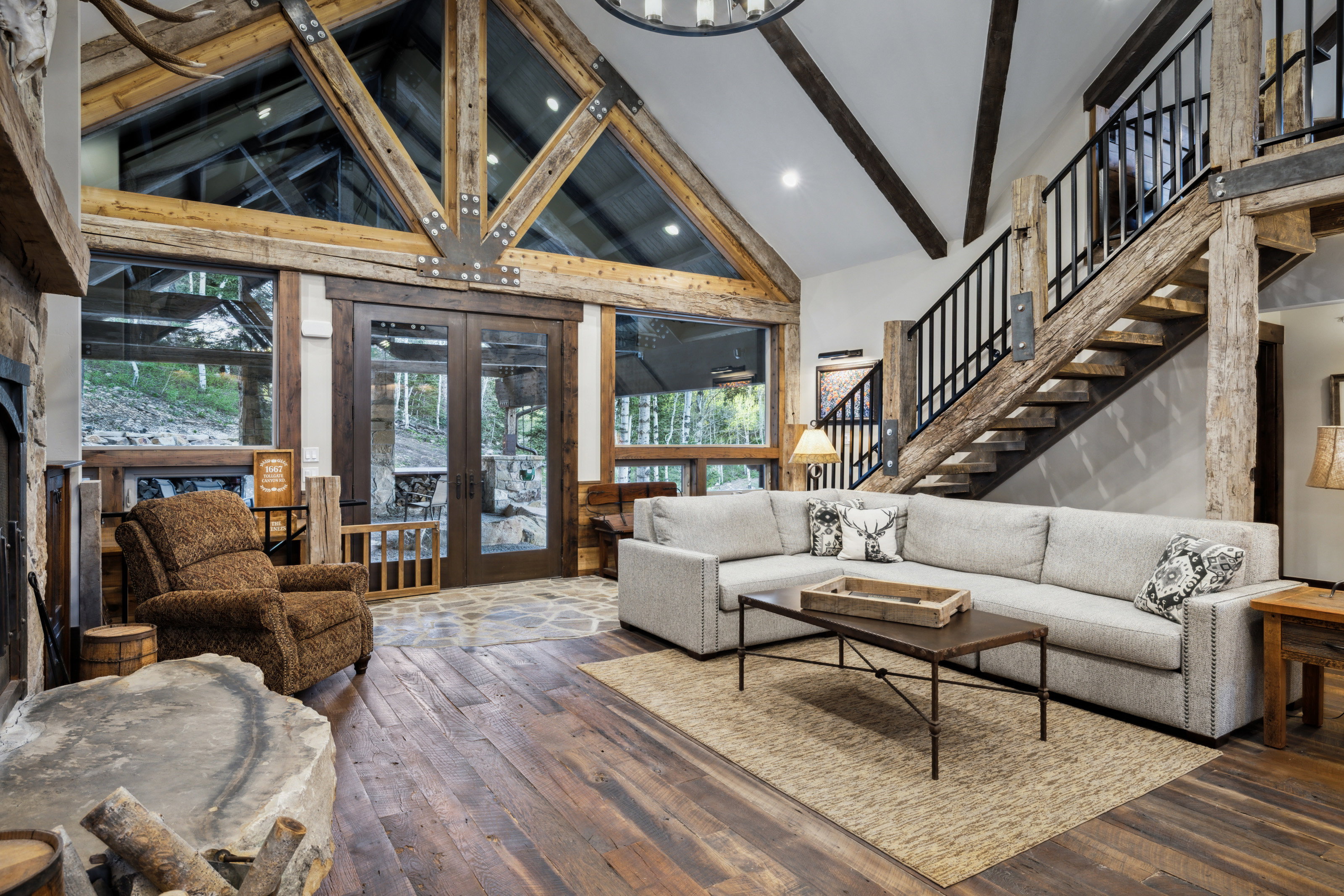 Vaulted ceiling with exposed wood beams and architectural glass, rich wood-finish flooring, stone fireplace, and a striking open staircase with iron railings