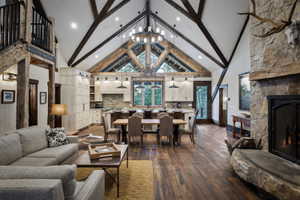 Open-concept living space featuring vaulted ceilings with exposed wood beams, wood-finish flooring, a stone fireplace with a substantial hearth, and a kitchen with light wood cabinetry and pendant lighting