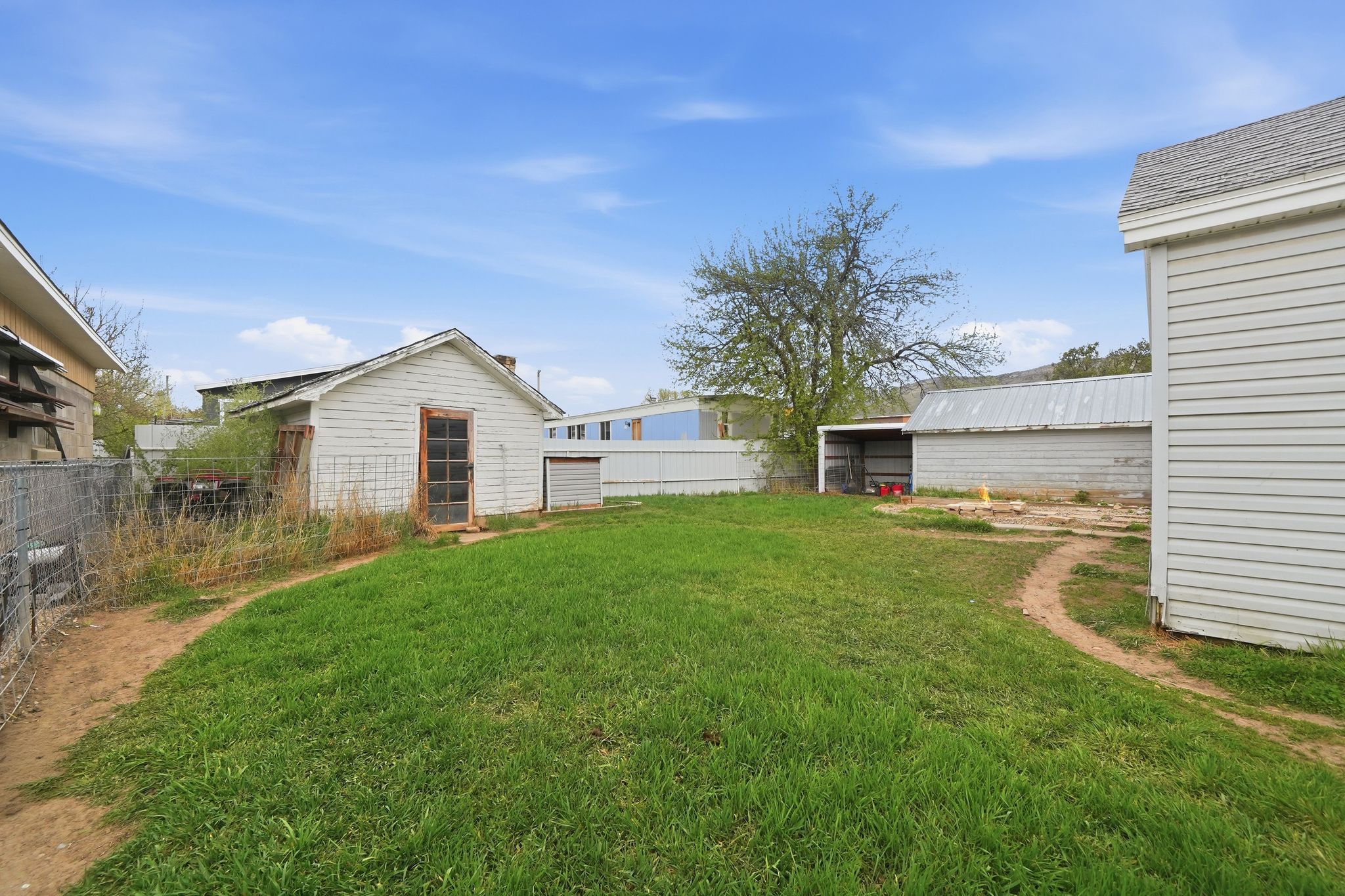Expansive green lawn with a white detached storage building, a metal fence, a bare earth path, and a partially covered metal structure