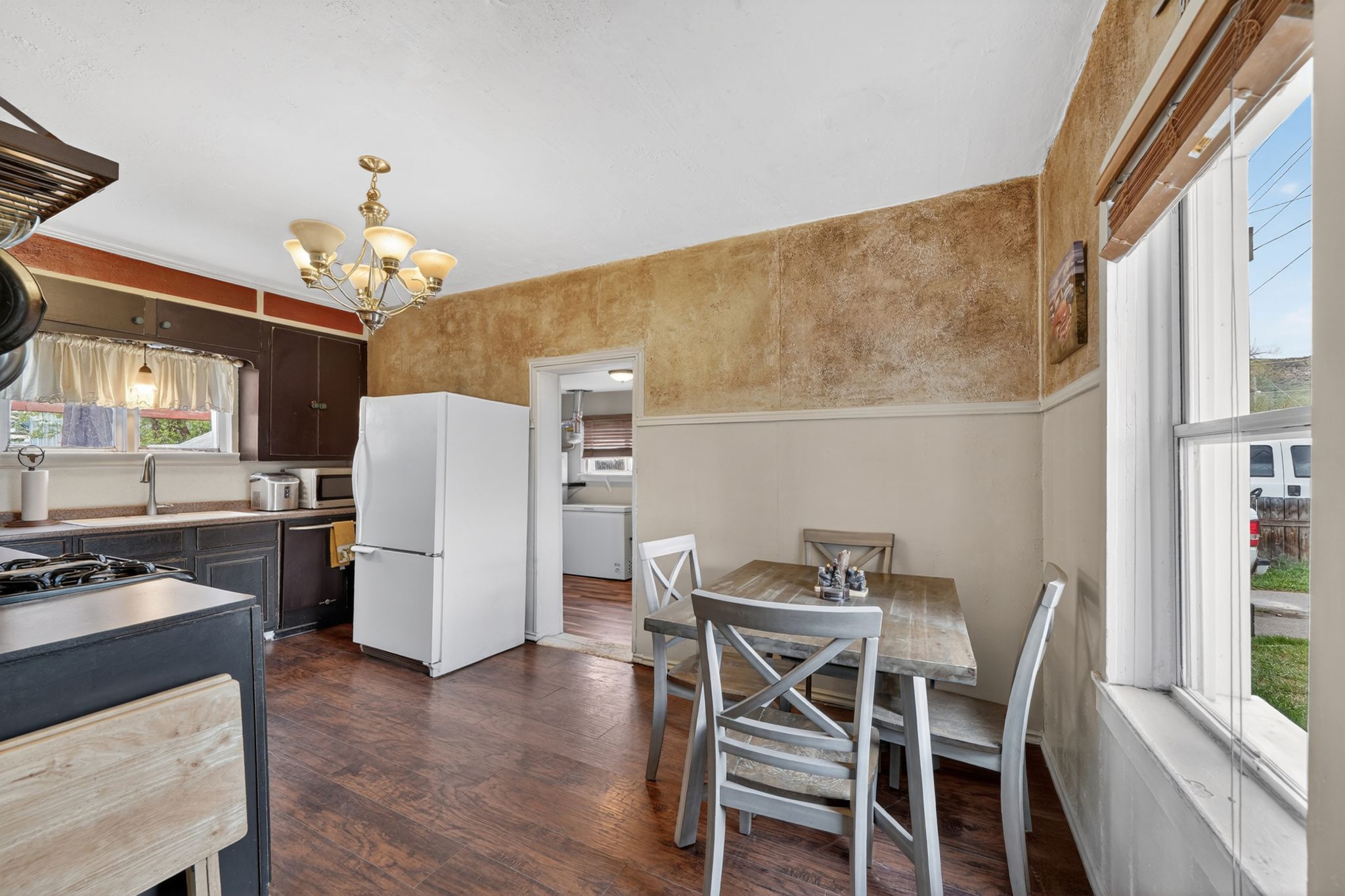 Kitchen dining area featuring wood-finish flooring, dark wood cabinetry, a four-light chandelier, a white refrigerator, and a window