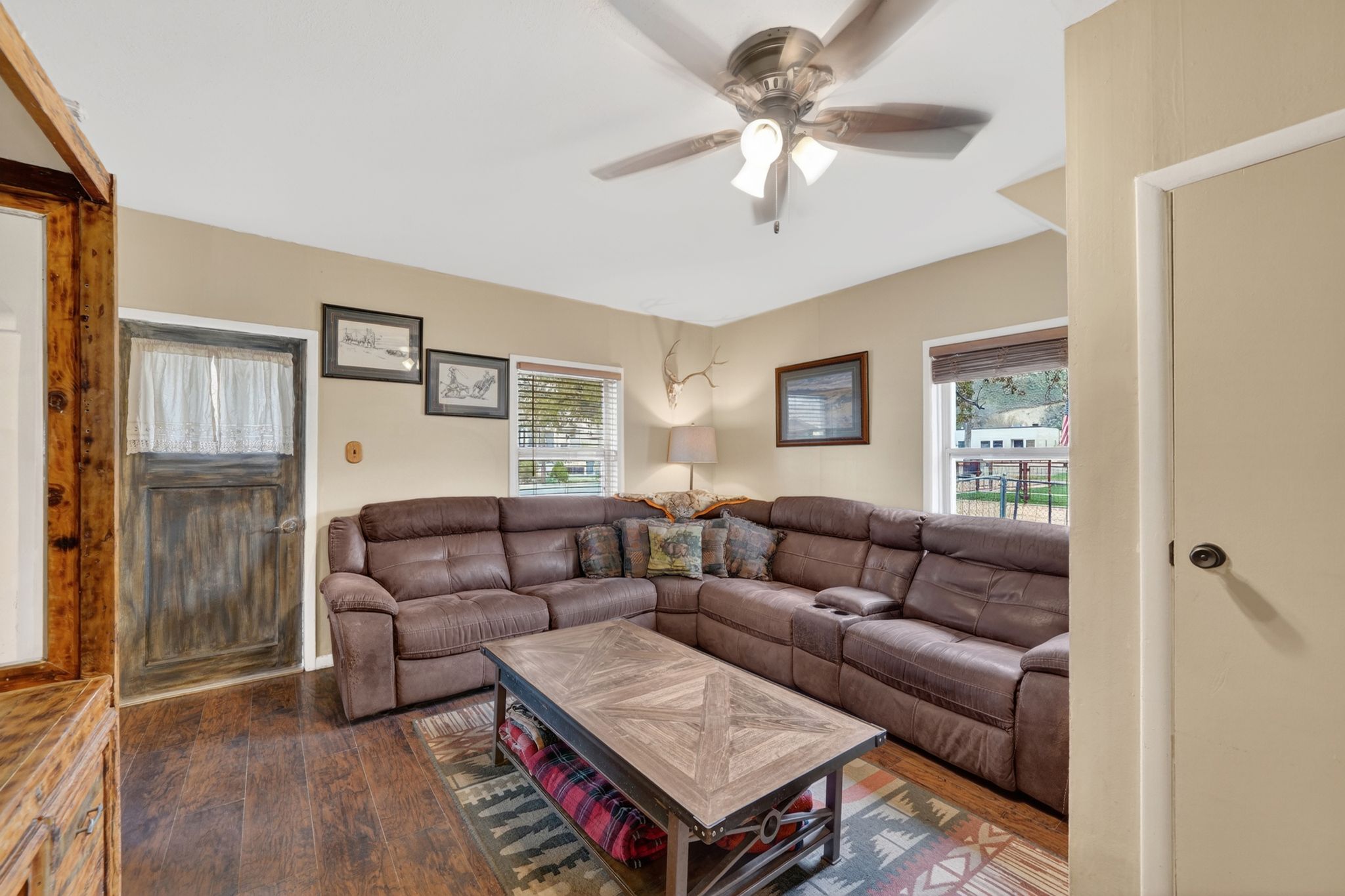 Living area featuring wood-finish flooring, neutral wall tones, a ceiling fan with integrated lighting, and multiple windows with blinds