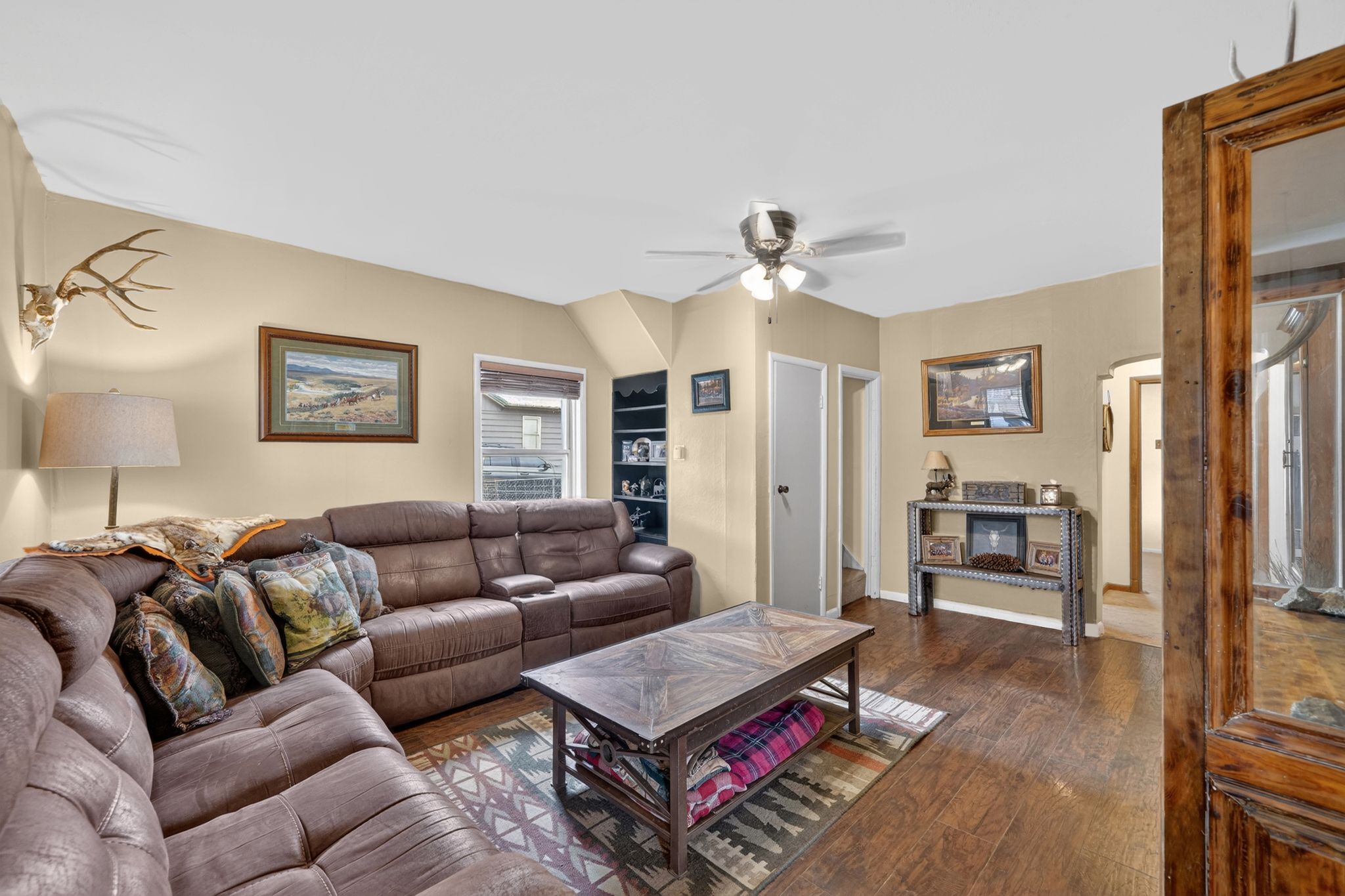 Living area featuring wood-finish flooring, light-toned walls, a ceiling fan with integrated lighting, a built-in shelving unit, and multiple windows providing natural light