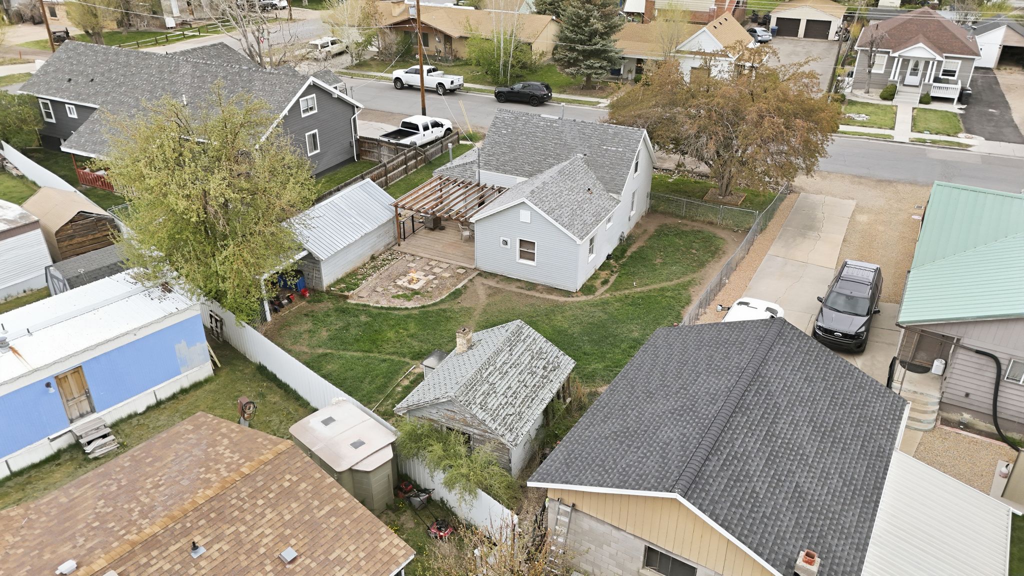 Aerial perspective showcasing the property with a light gray exterior, a detached garage, a fenced yard, a pergola-covered patio, and a fire pit area