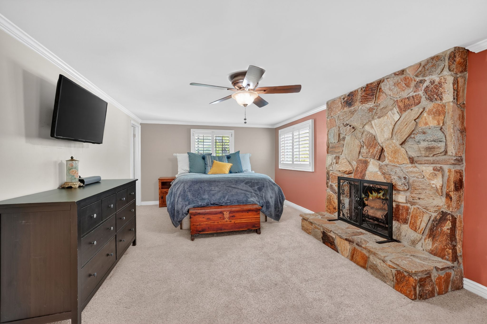 Spacious bedroom featuring a natural stone fireplace, ceiling fan, carpet flooring, and plantation shutters