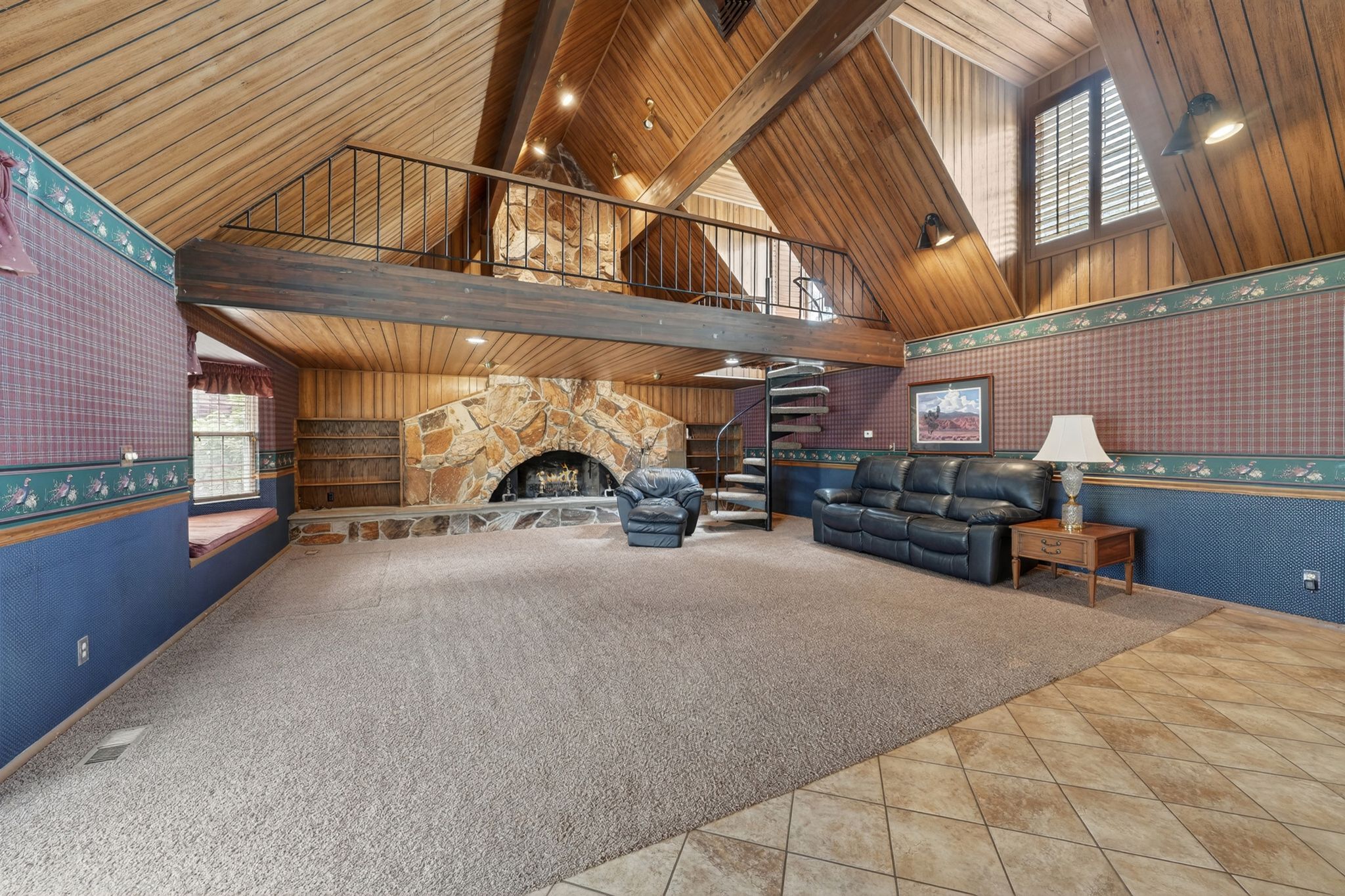 Vaulted wood-paneled ceiling with exposed beams, stone fireplace, built-in shelving, spiral staircase, and an upper-level loft with a metal railing