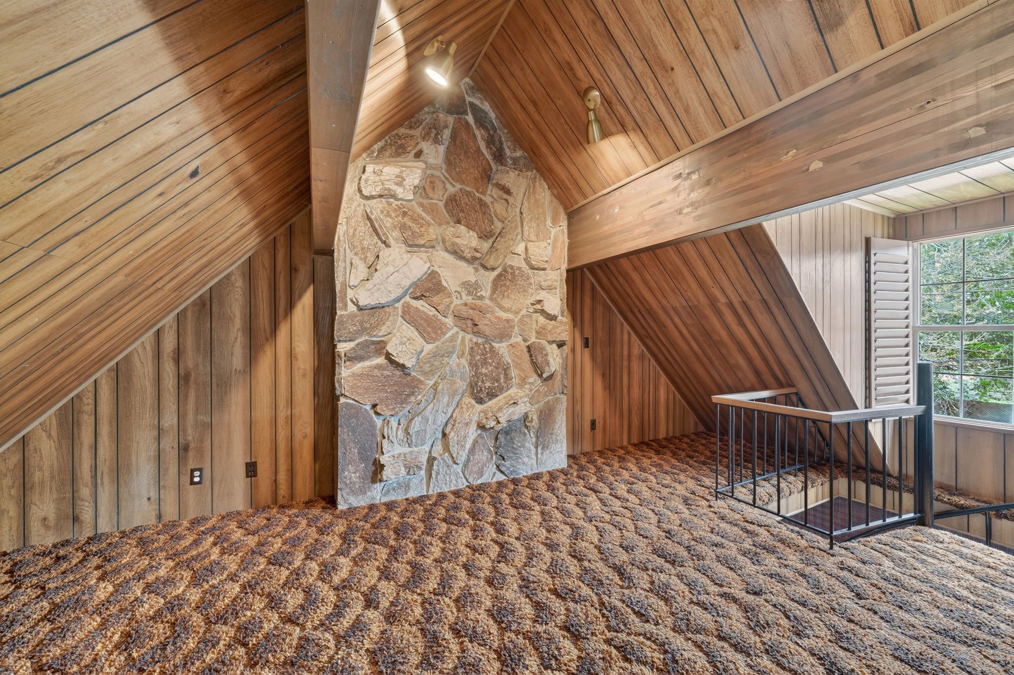 Loft space featuring a prominent stone fireplace, vaulted wood-paneled ceilings, textured carpeting, and a window with white louvered shutters overlooking exterior trees
