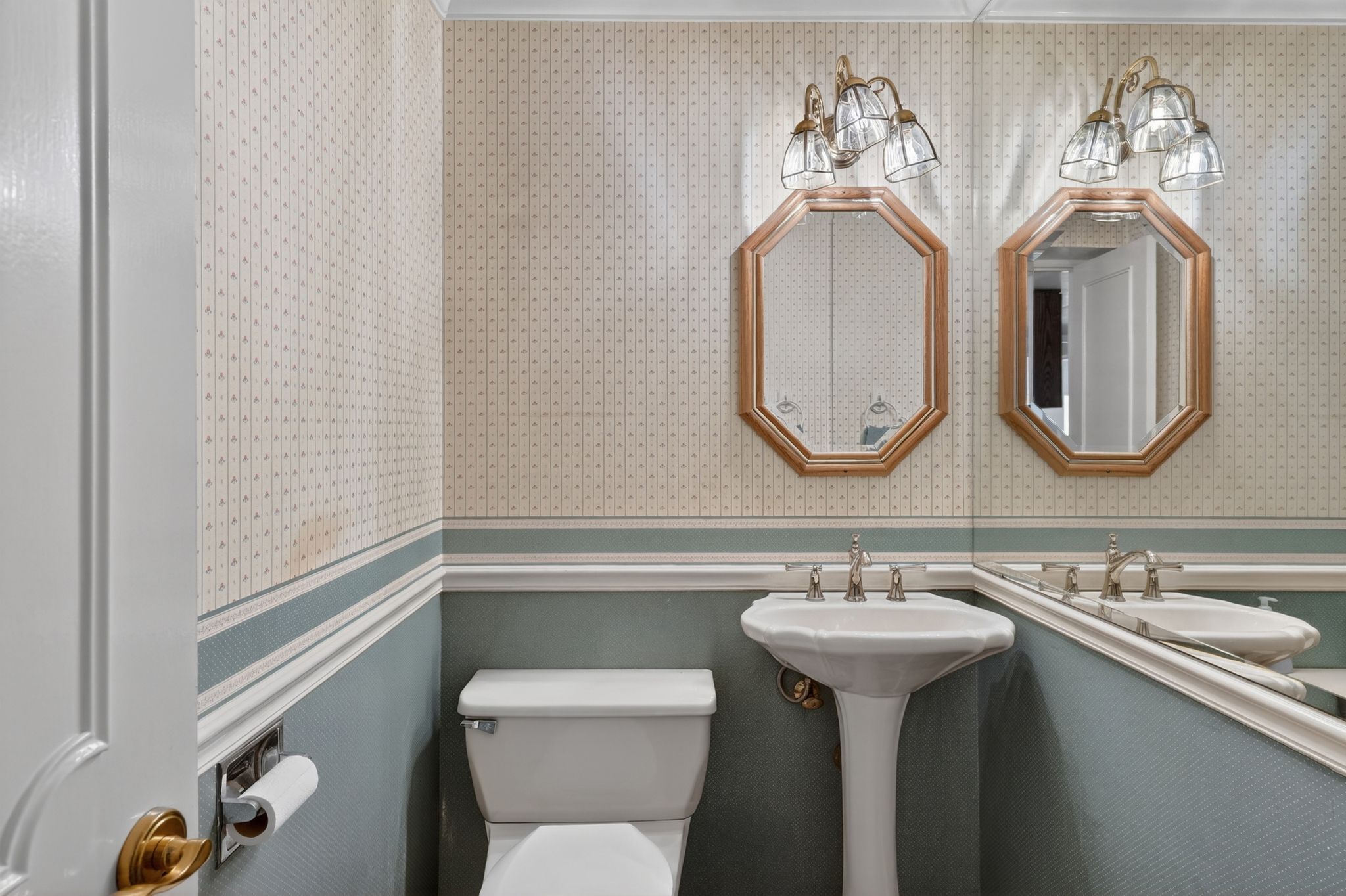 Pedestal sink with brass-finish faucet, octagonal mirrors with brass-finish frames, wall-mounted lighting fixtures, full wall mirror, and patterned wallpaper