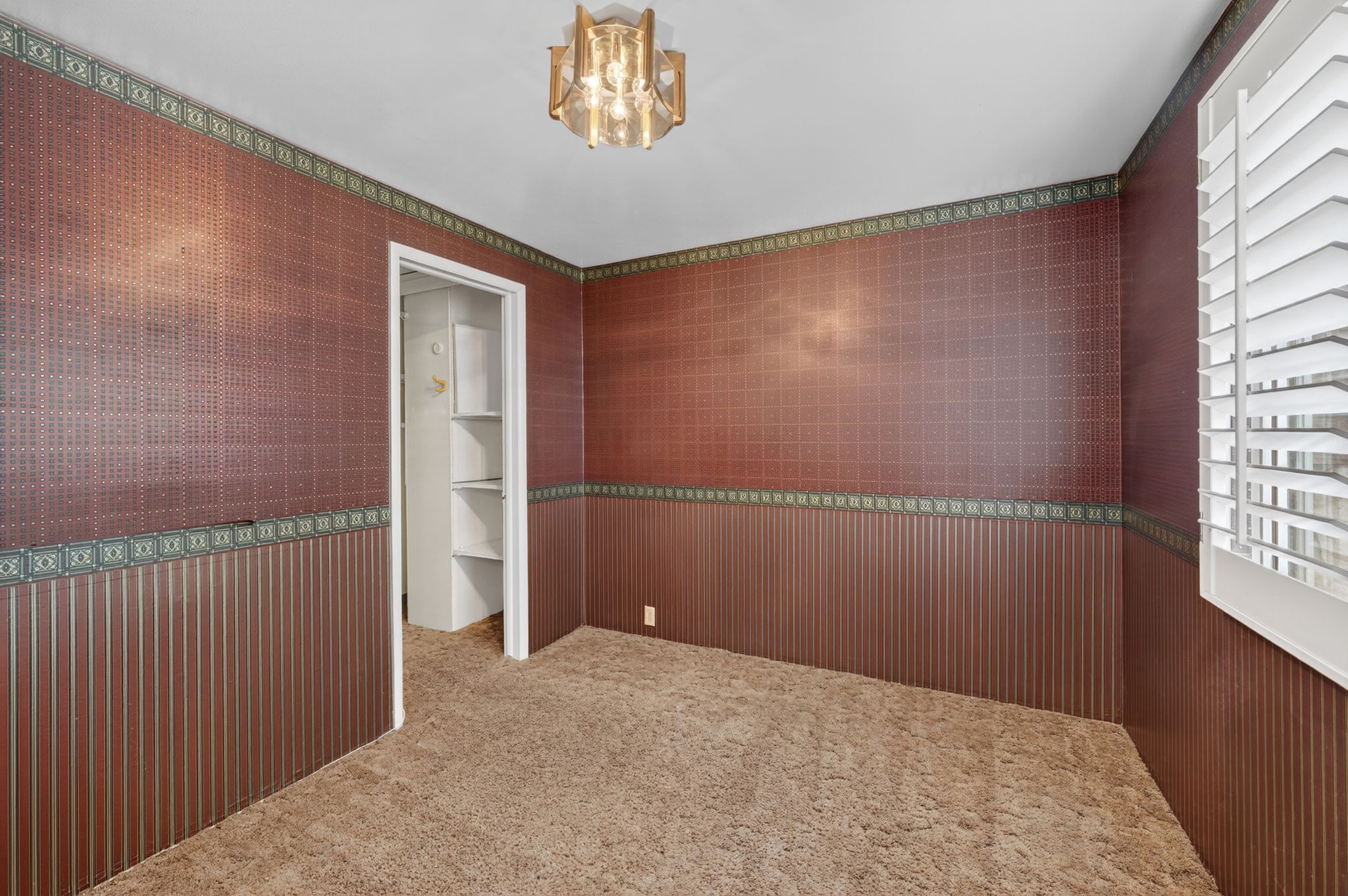 Room featuring a textured brown wallpaper with patterned accents, a ceiling-mounted light fixture, and a window with white plantation shutters