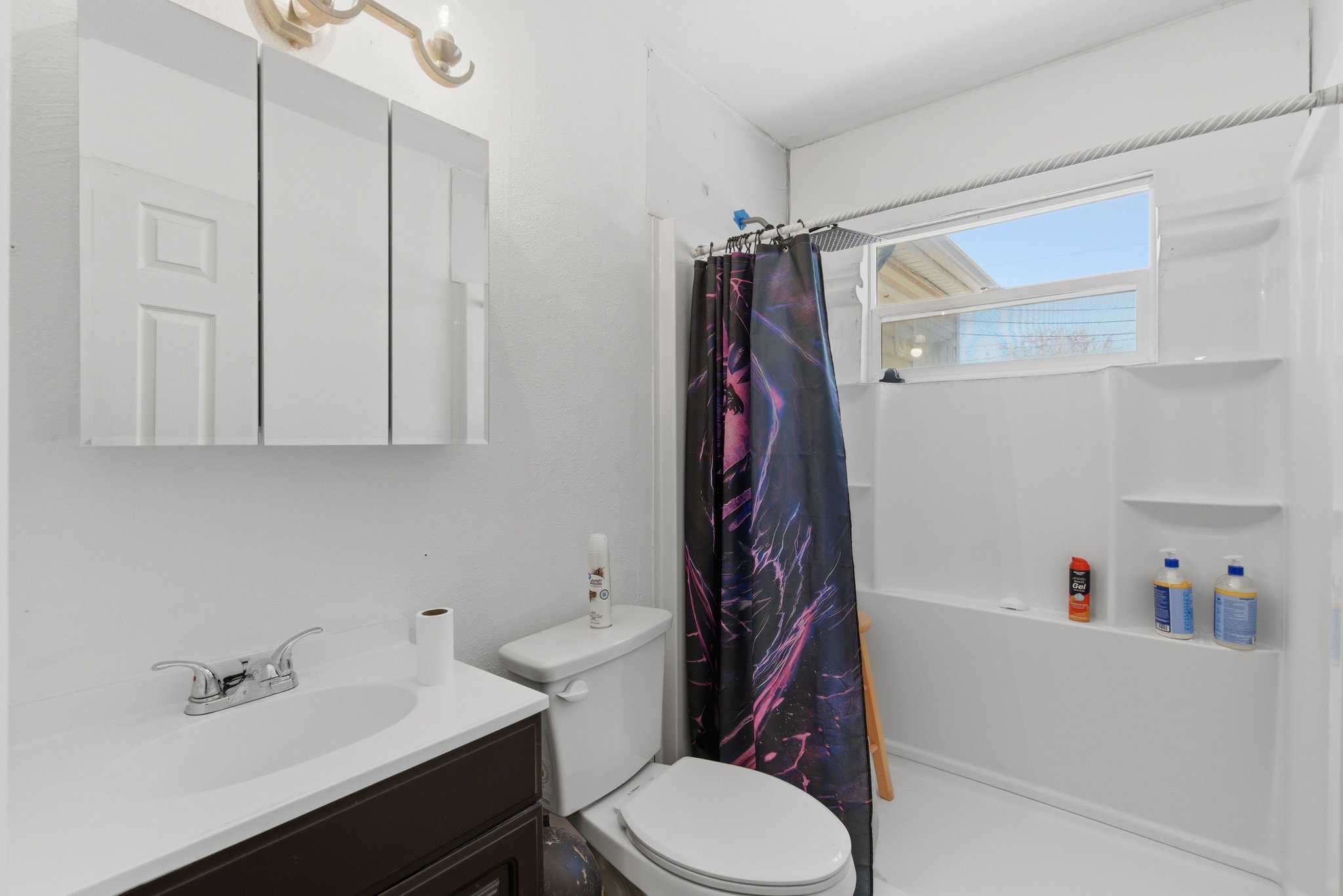 Bathroom featuring a white vanity with a dark wood-finish cabinet, a mirrored medicine cabinet, a toilet, a shower/tub combination with a window, and built-in shelving within the shower surround
